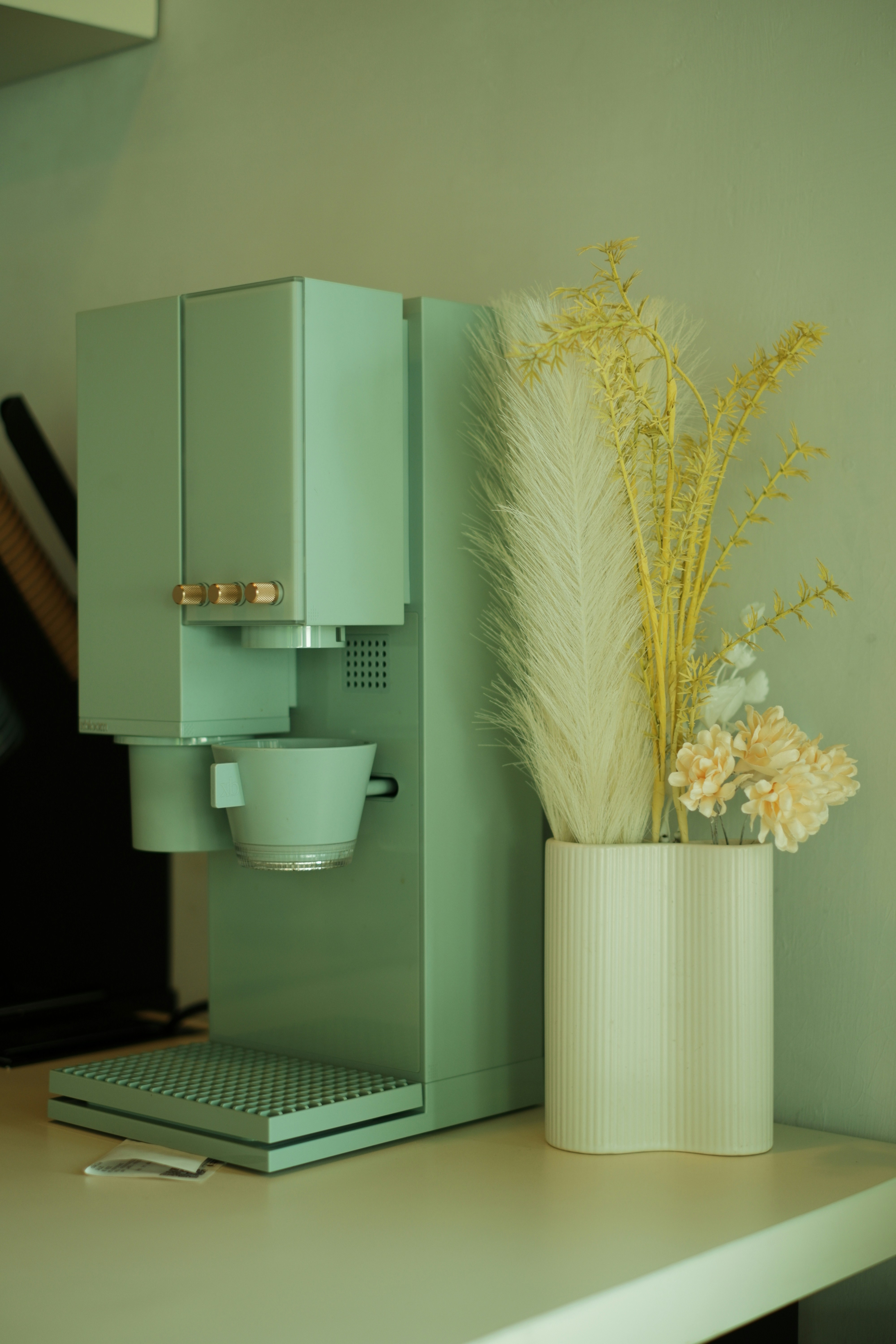 A 3D render of a mint green coffee machine beside a white vase with dried flowers
