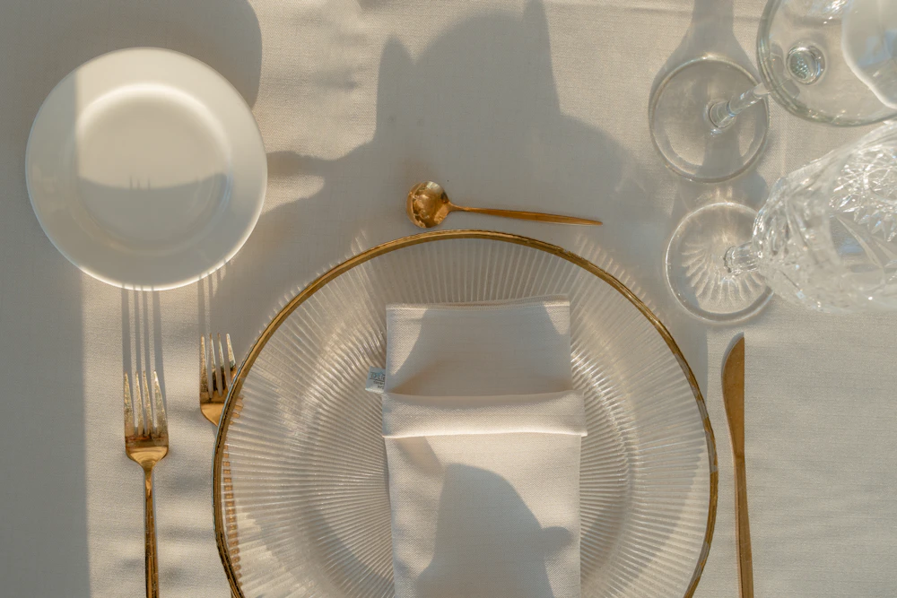 A minimal place setting with a glass plate, gold cutlery and folded white napkin