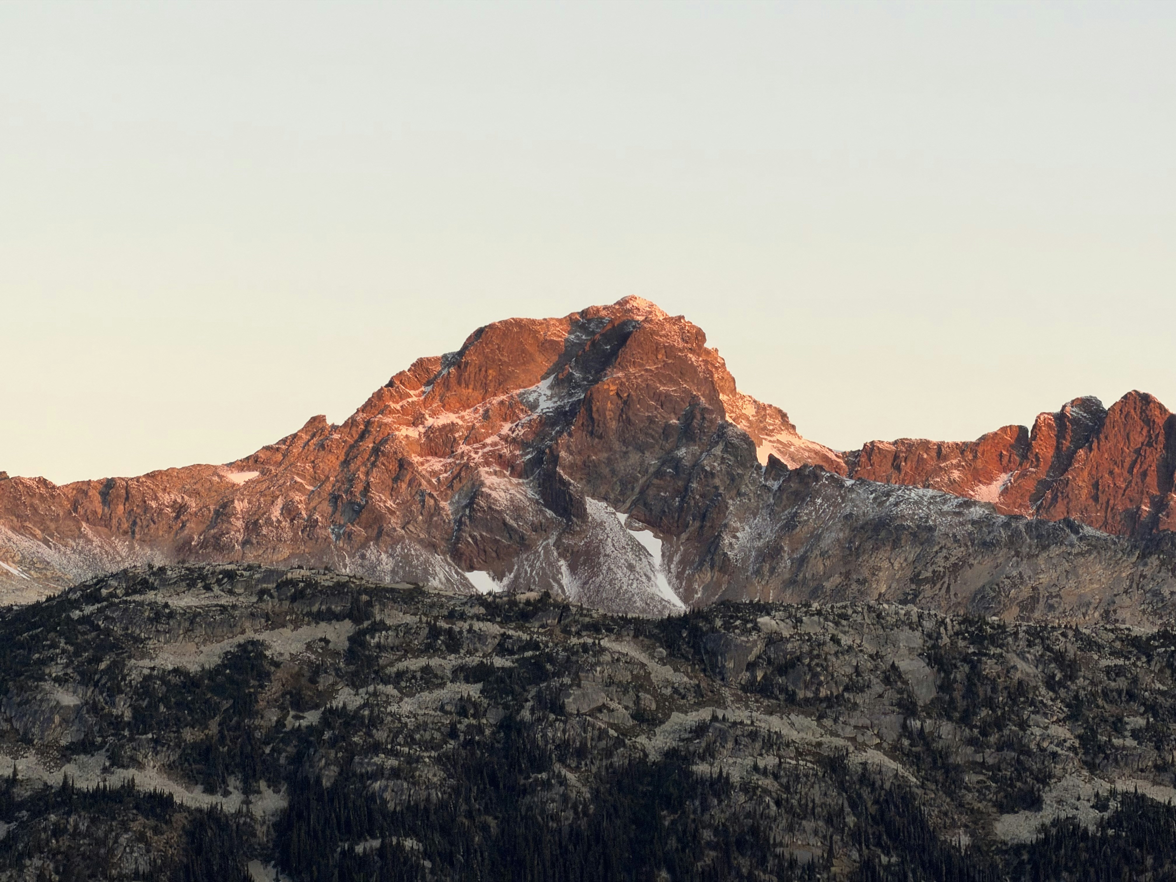 A snow-dusted mountain peak illuminated by warm golden hour light above a dark forested slope