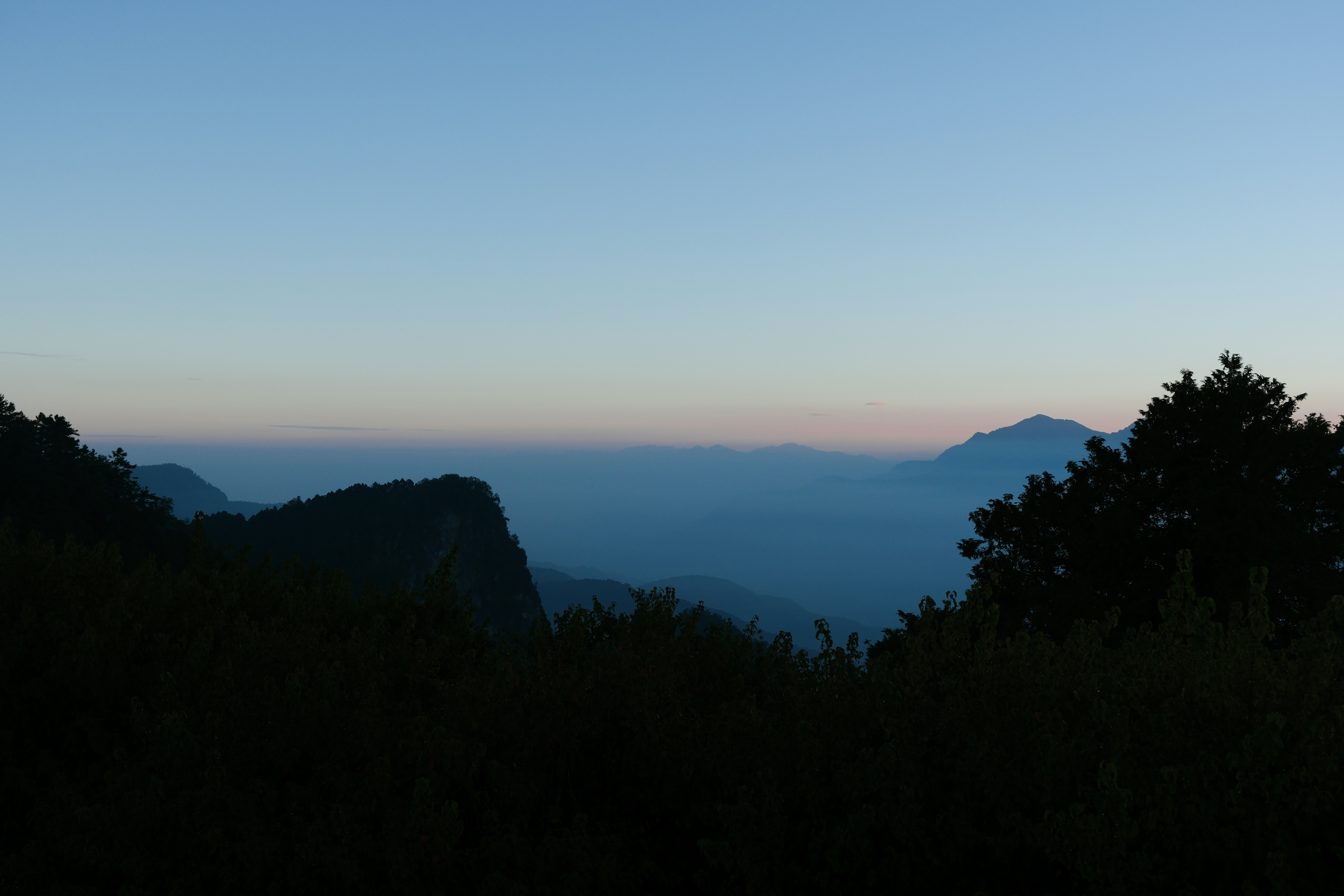 Árboles en silueta enmarcan una vista lejana de crestas montañosas azules superpuestas con poca luz
