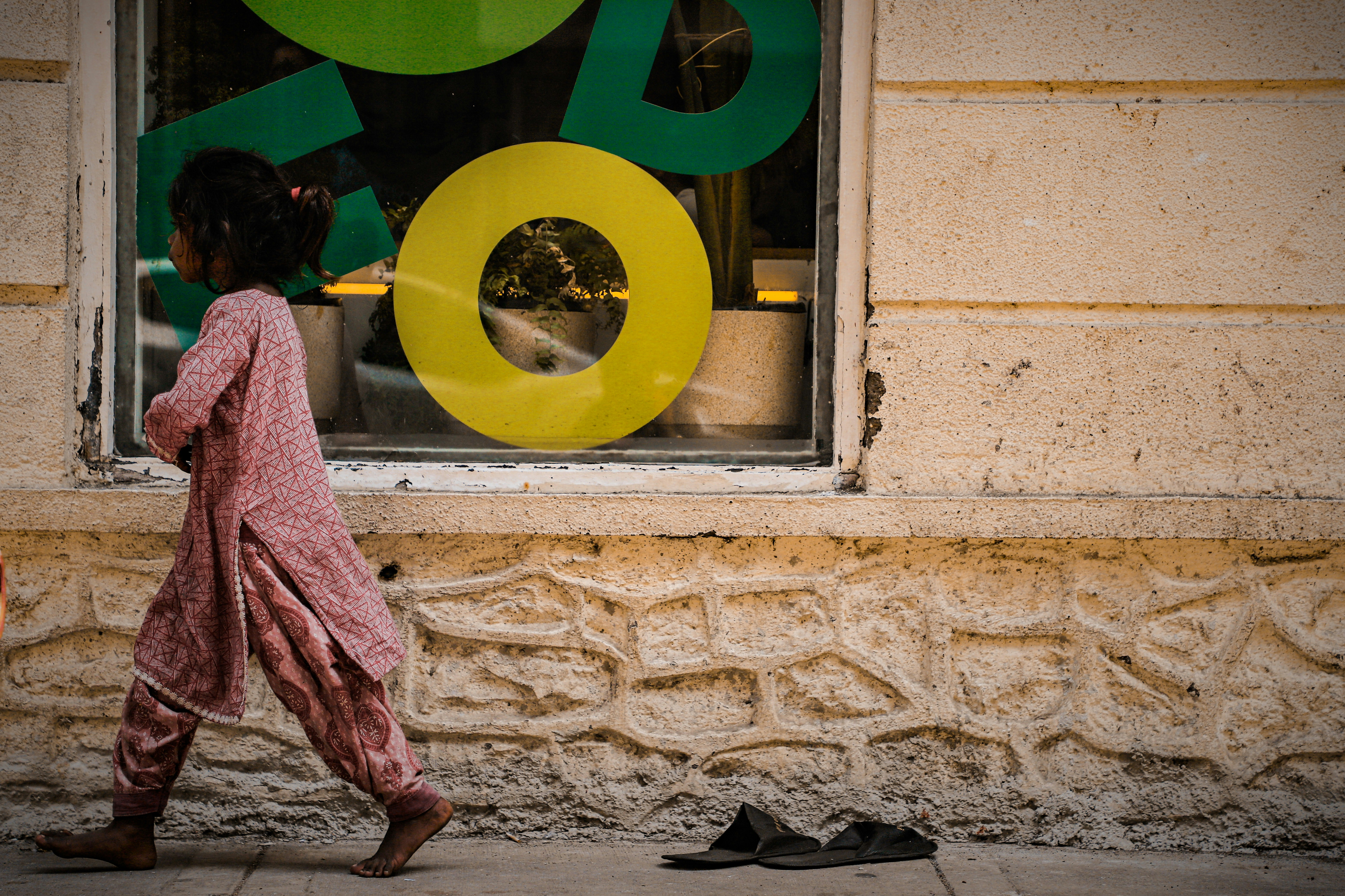 A barefoot child in a patterned pink tunic walking past a shop window with large yellow lettering