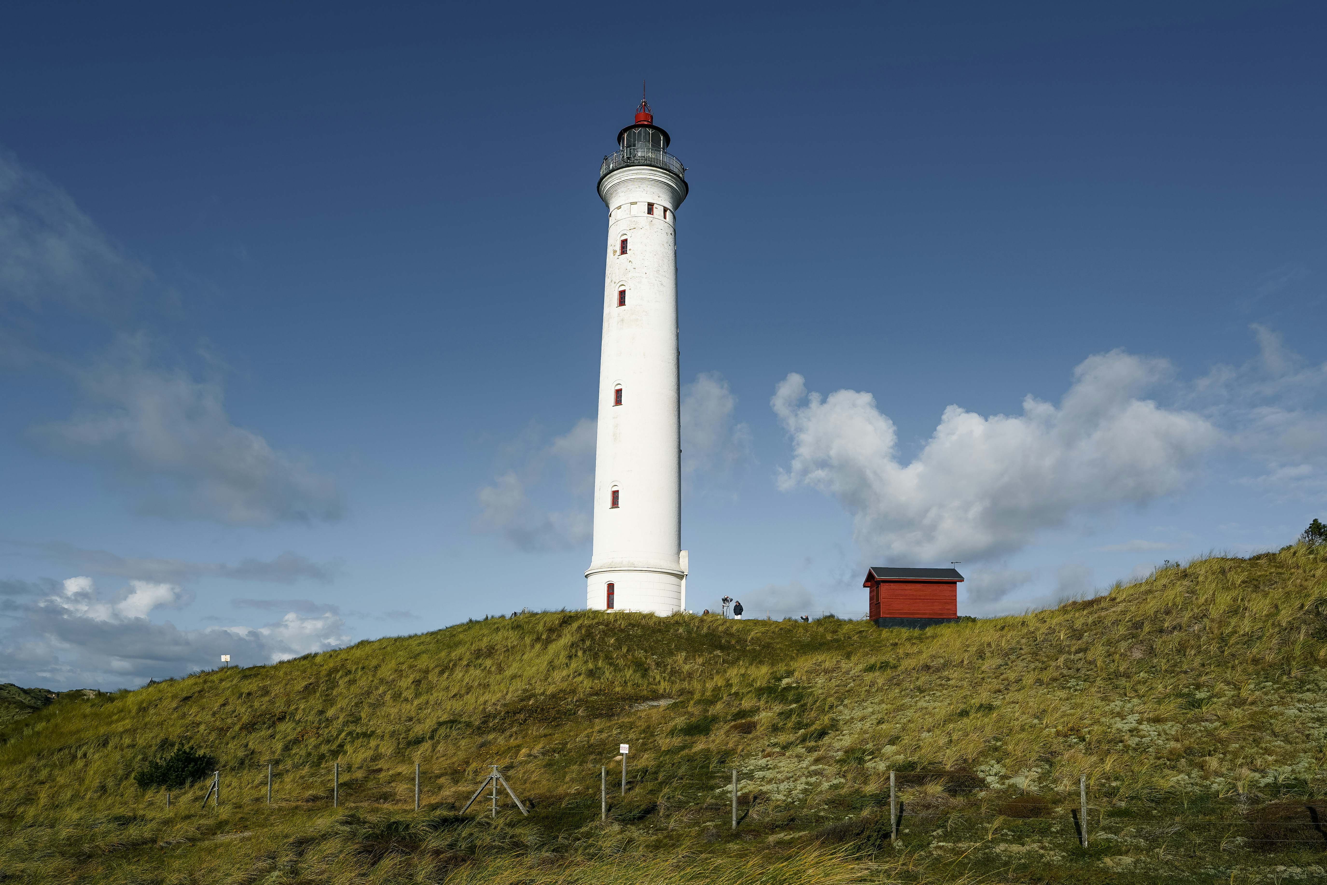 A tall white lighthouse on a grassy dune under a clear blue sky
