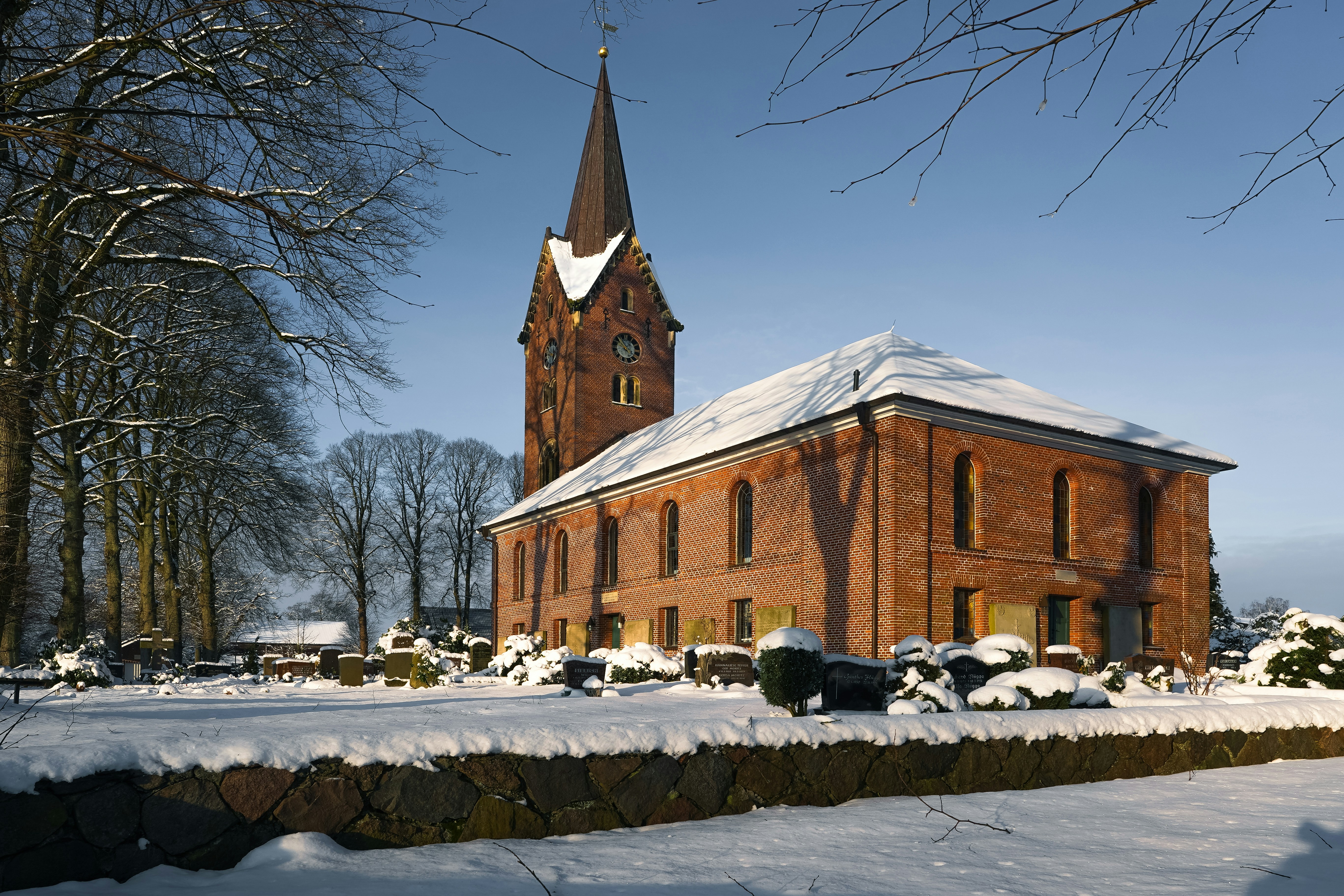 A red brick church with a tall steeple in a snow-covered cemetery under a clear blue sky