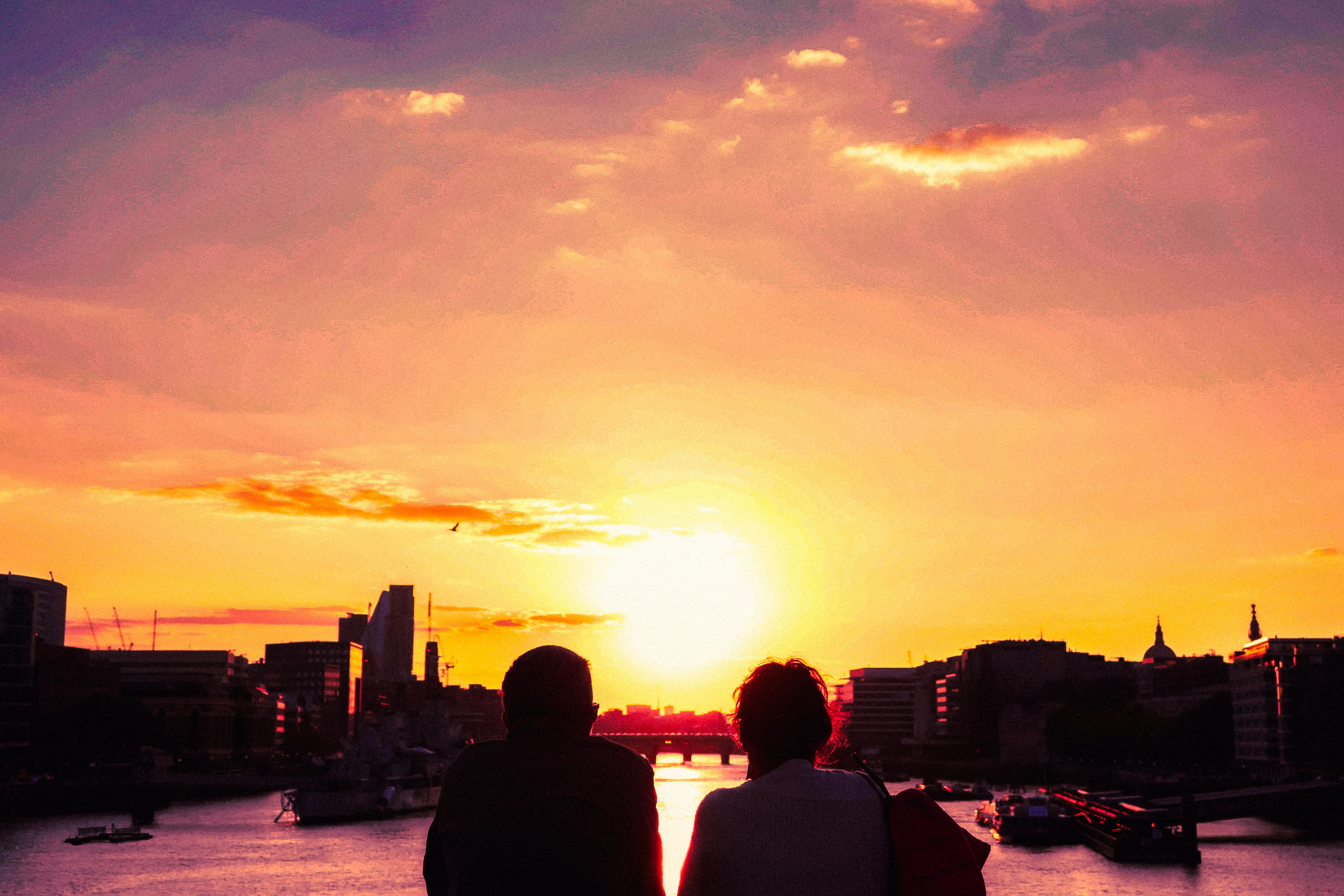 Two silhouetted figures watch the sunset over a city river, with warm oranges and pinks reflecting on the water; a skyline and boats anchor the scene. (Photo / illustration-like sunset scene).