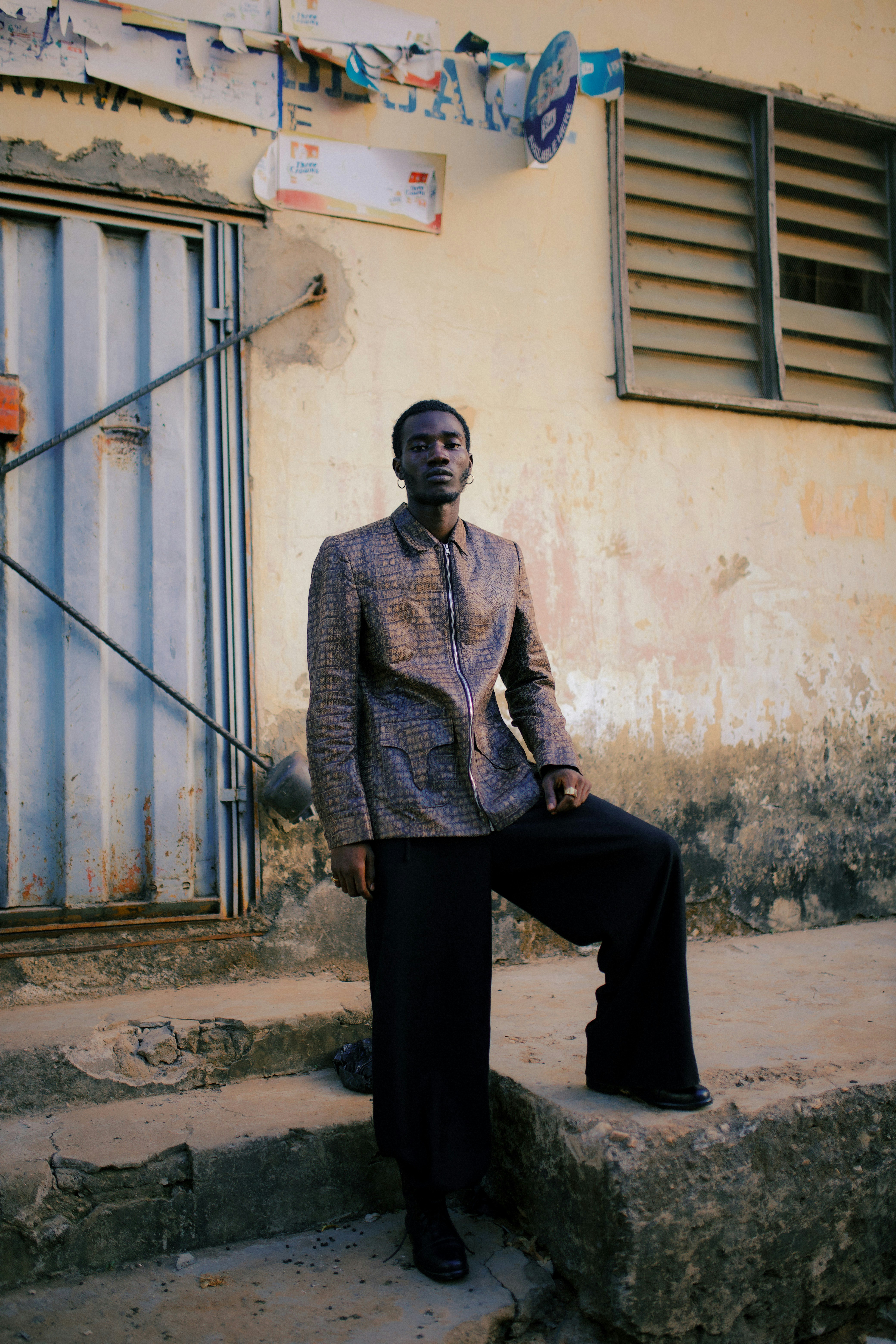 A man in a textured brown jacket and black trousers on concrete steps against a weathered wall