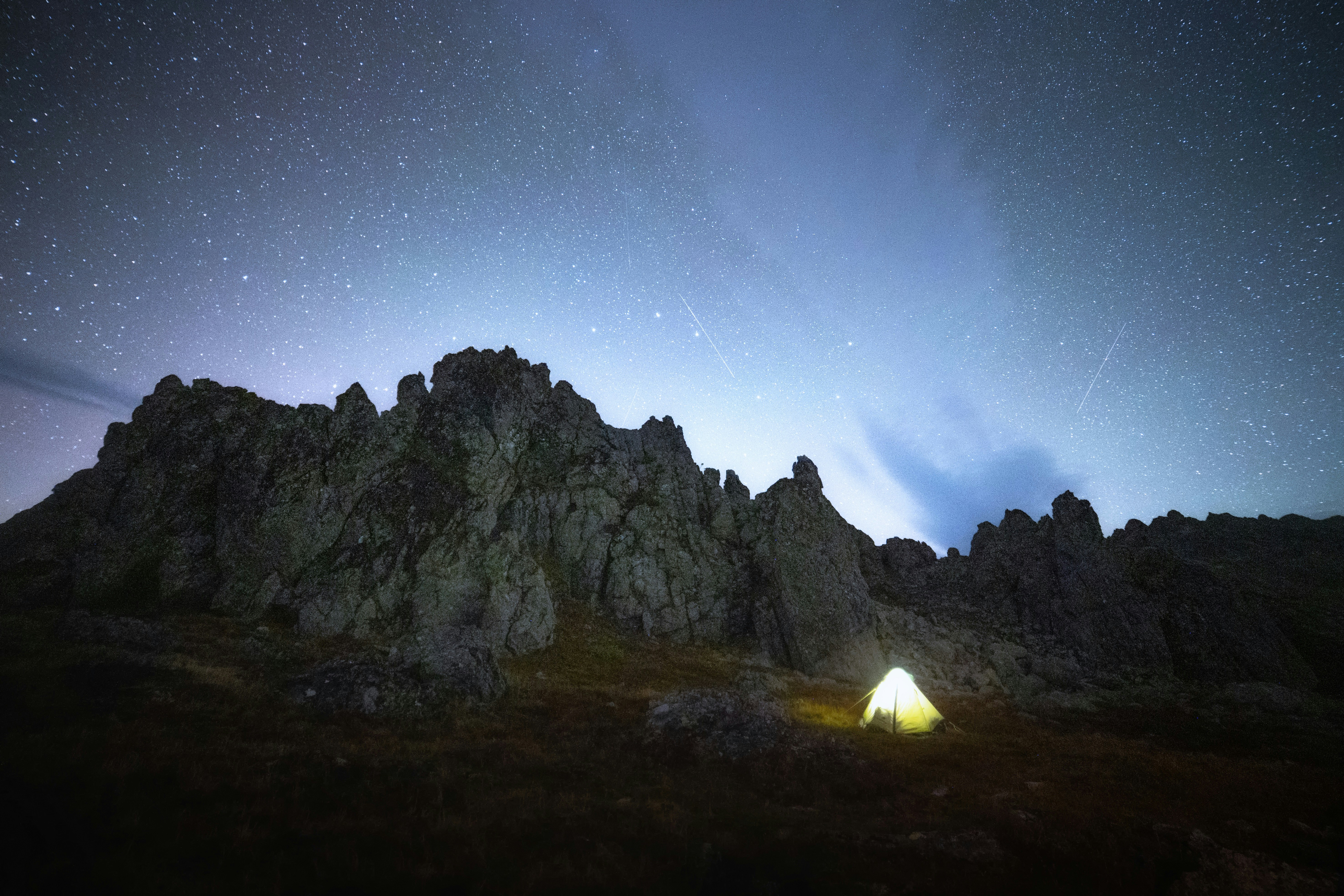 A glowing tent at the base of a rocky mountain under a starry night sky