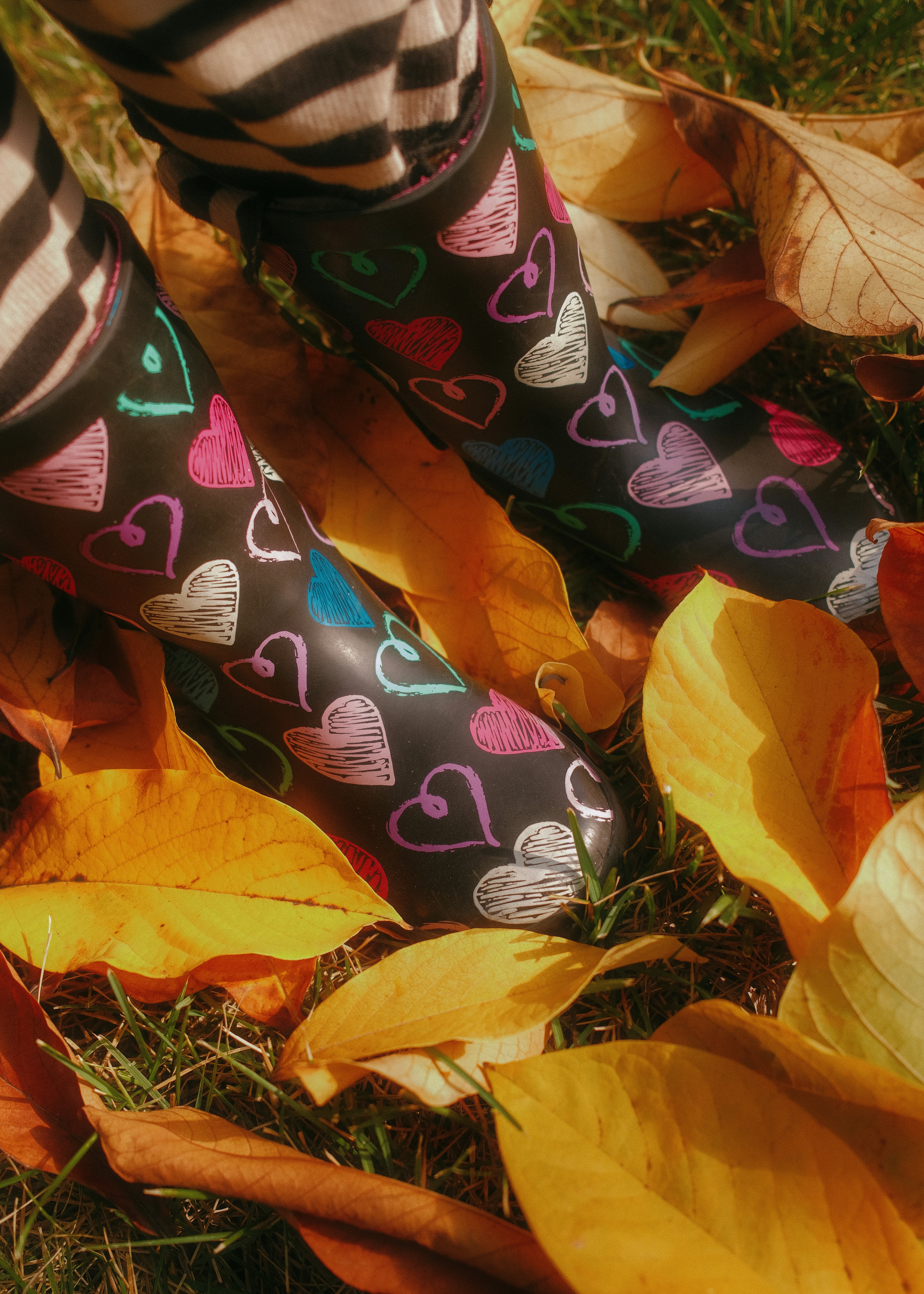 A child wearing black rain boots with colorful heart patterns standing in fallen autumn leaves