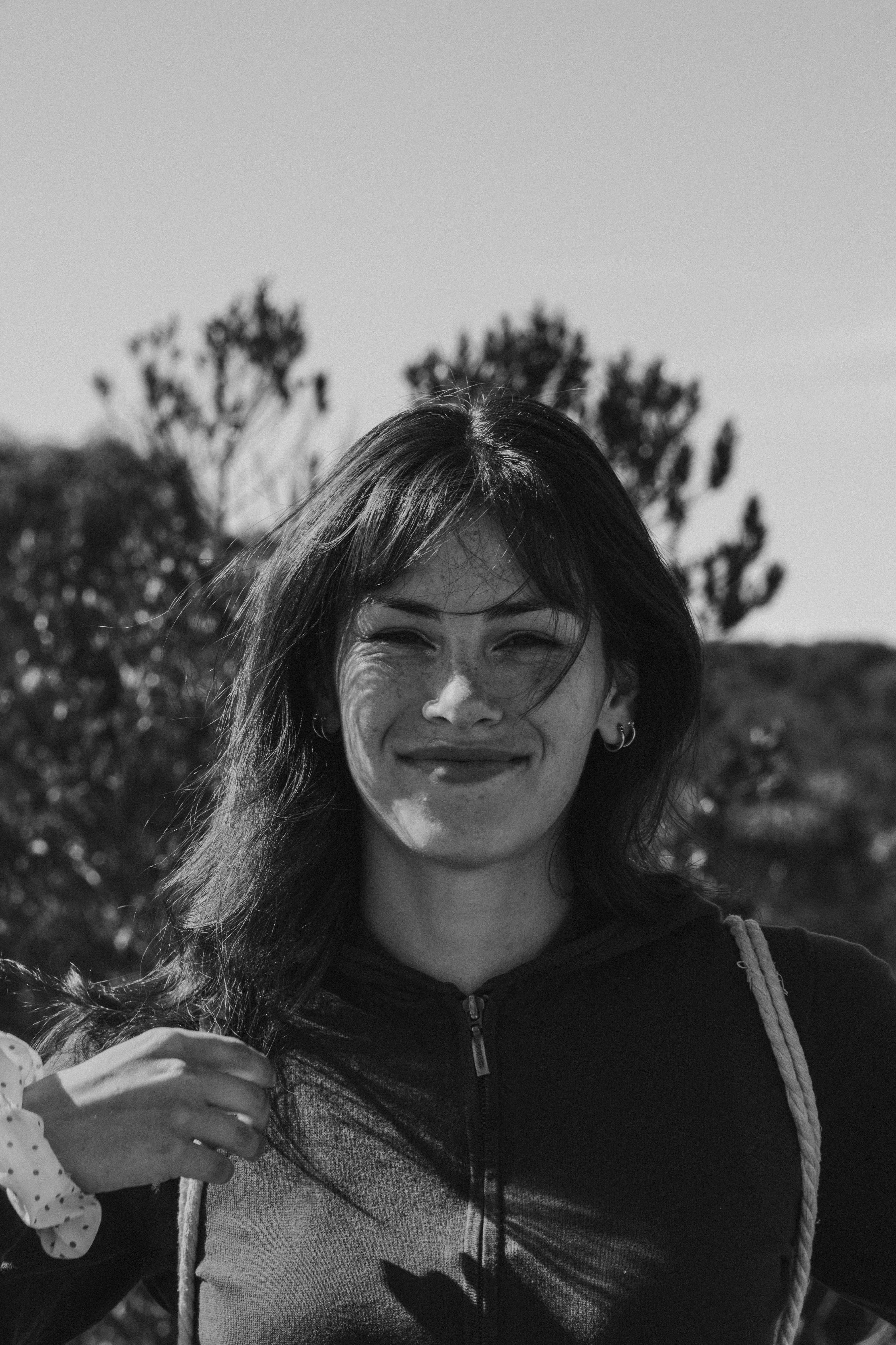 Uma mulher sorridente com franja e sardas usando um moletom preto com zíper ao ar livre