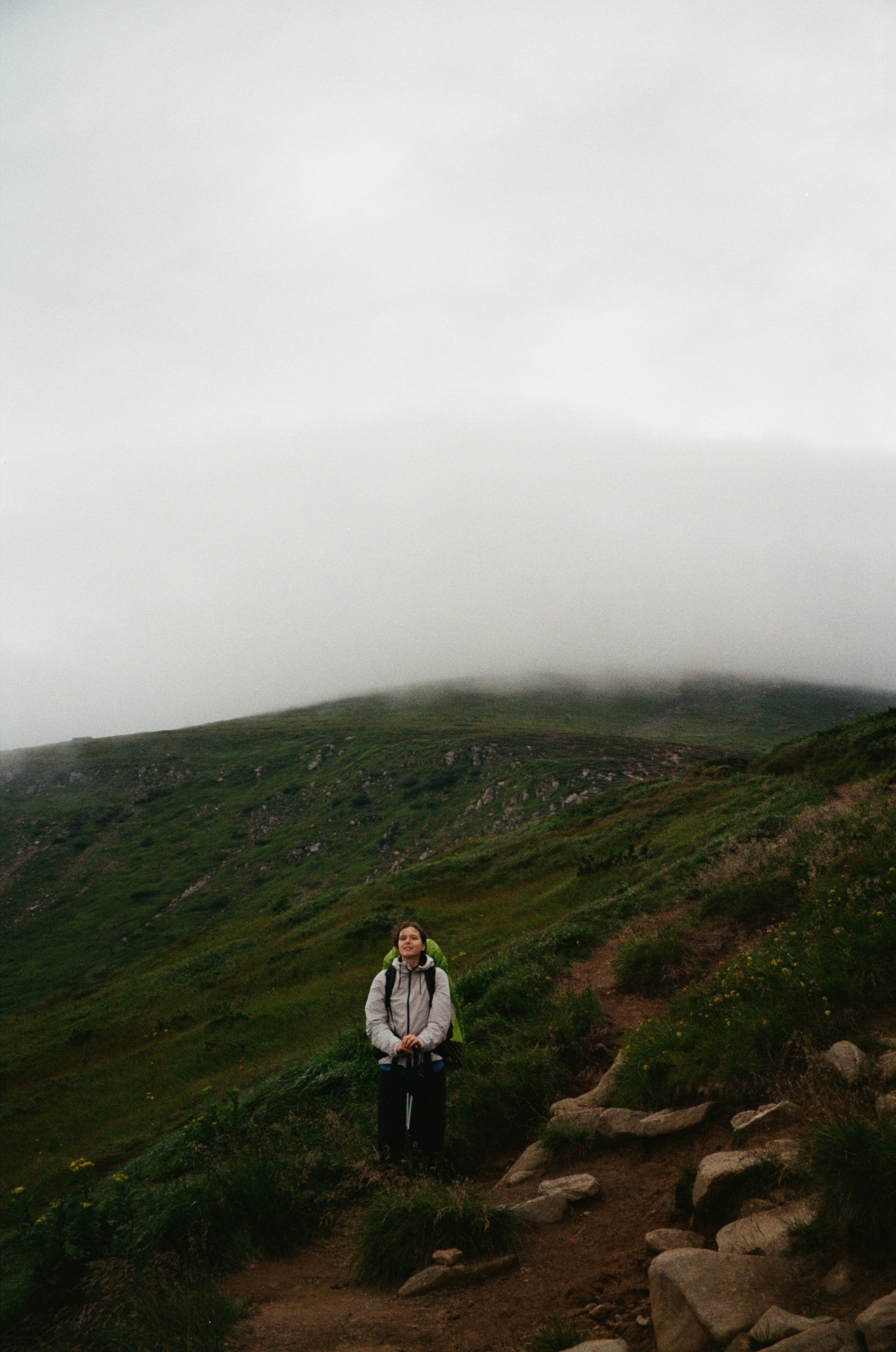 A person with a backpack on a grassy mountain trail under a thick layer of fog