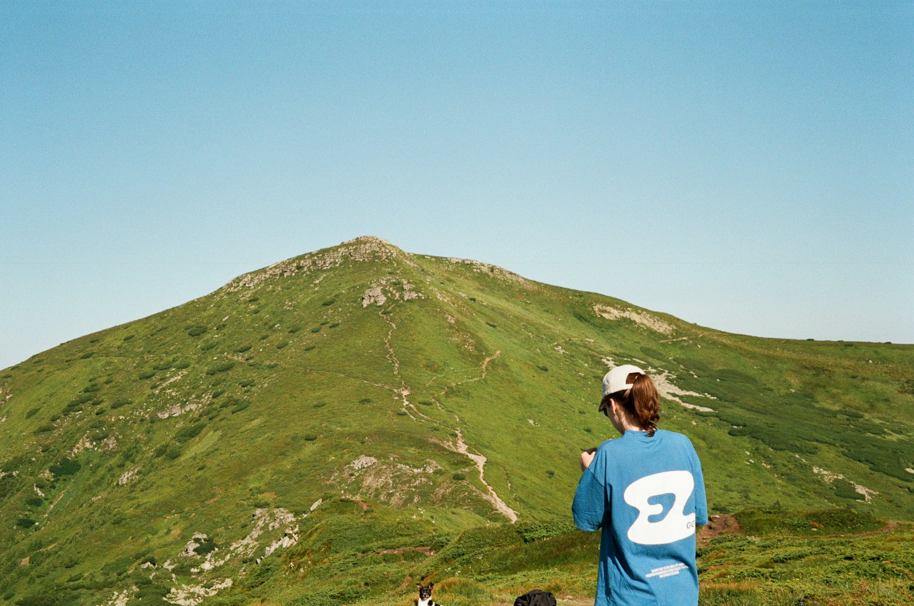 A person in a blue t-shirt and cap looking at a green mountain under a clear sky
