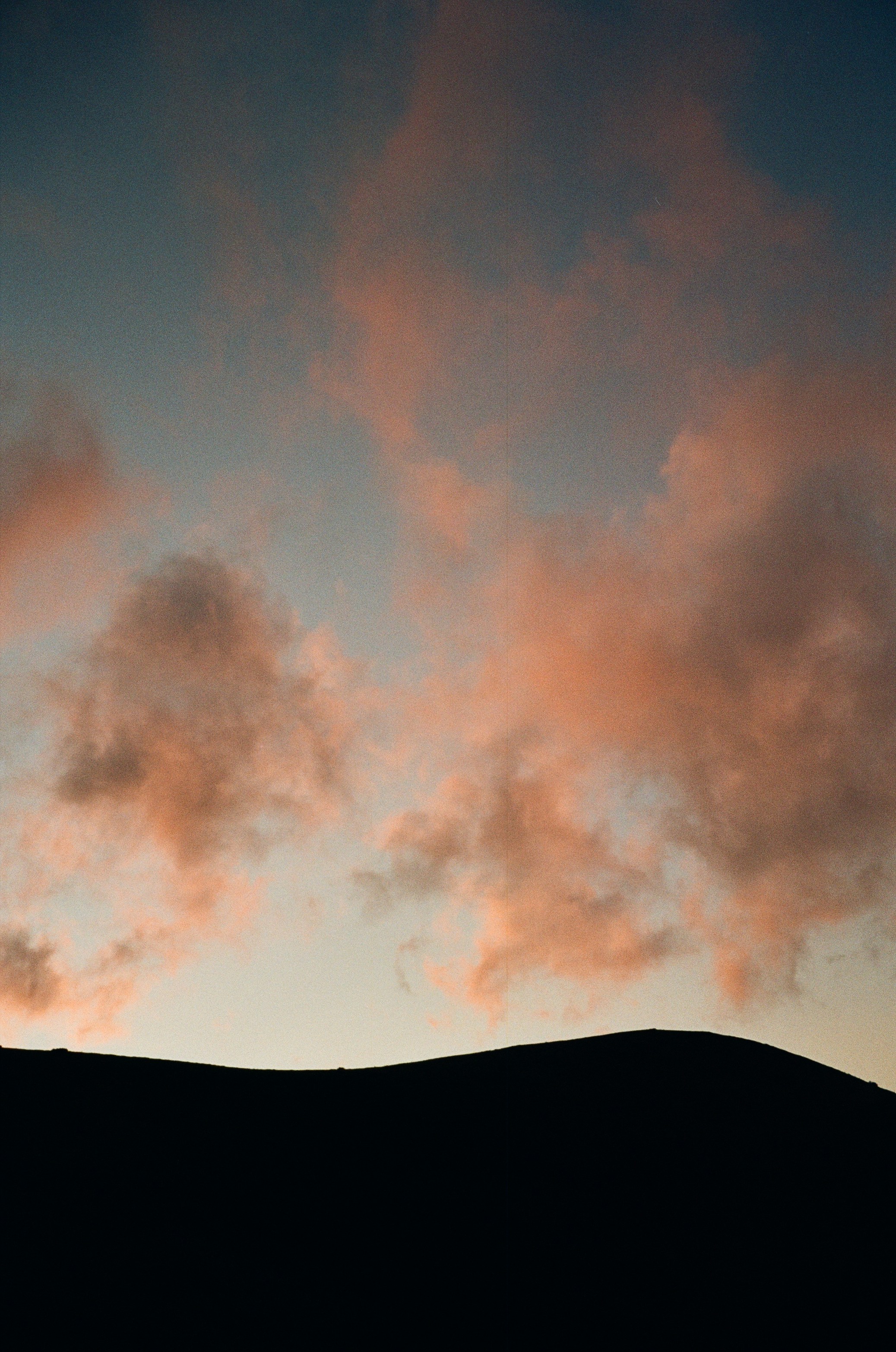 Soft pink clouds drifting over a dark mountain silhouette against a pale sky