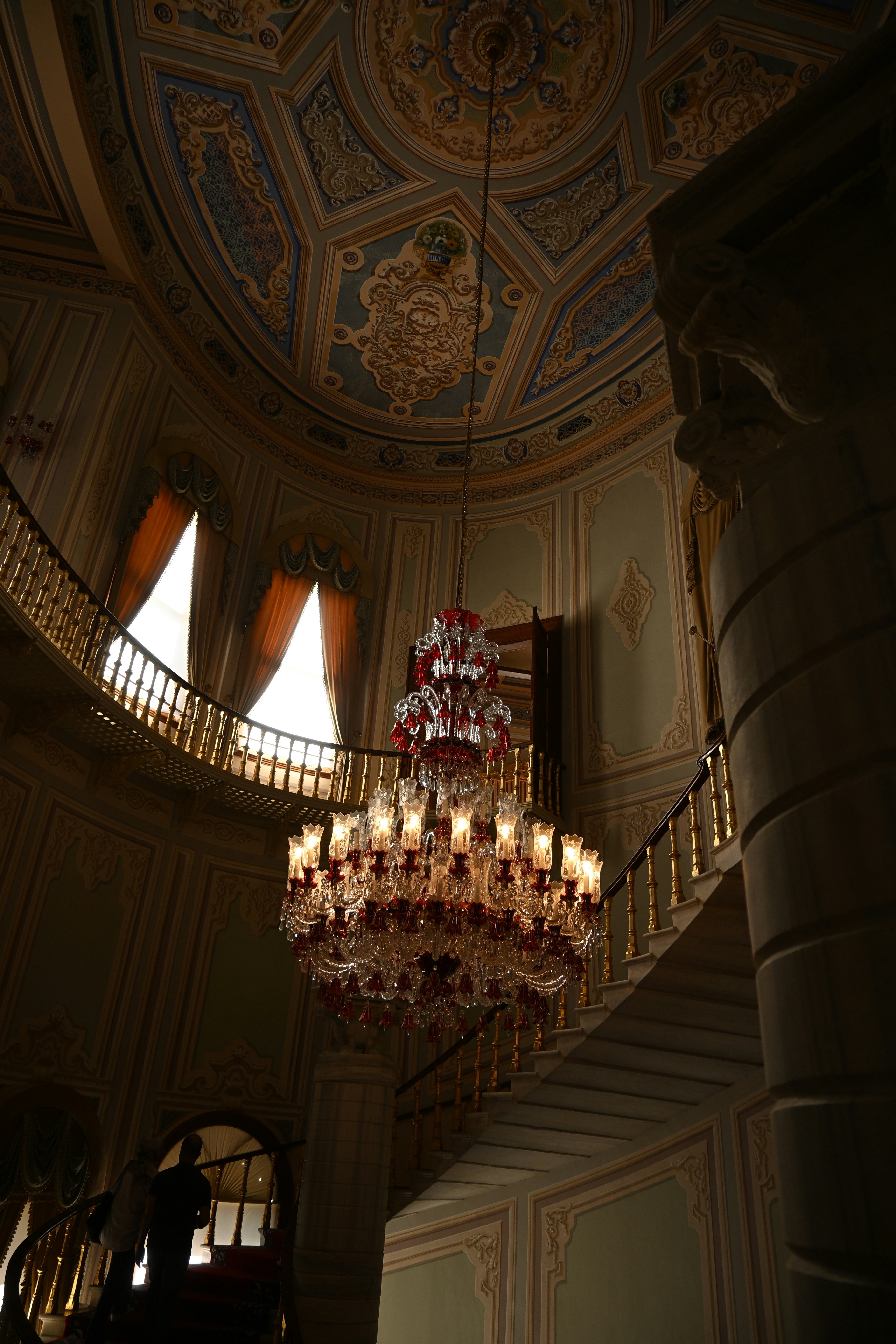 A large crystal chandelier hanging in a grand hall with an ornate ceiling and staircase