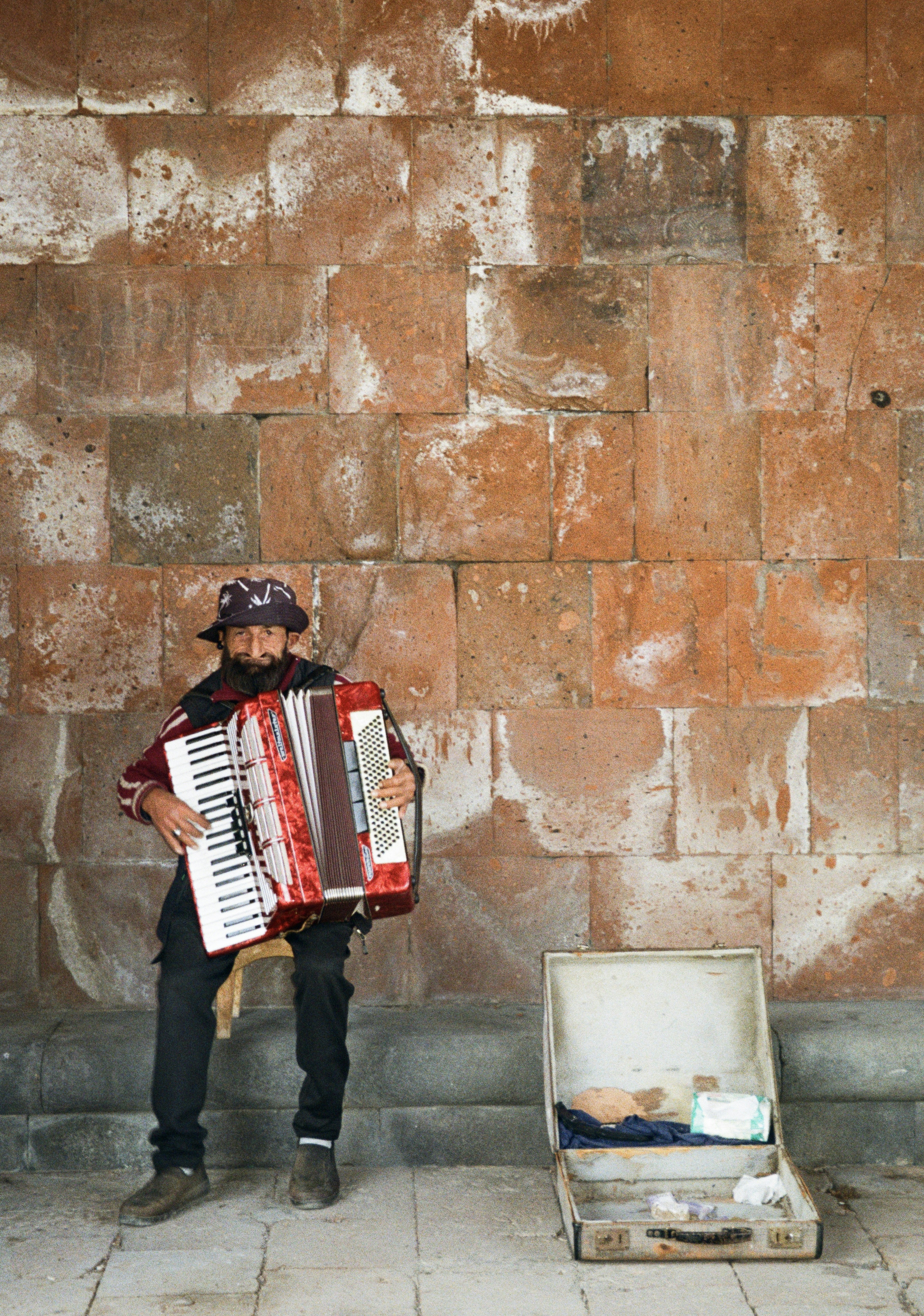 A bearded man in a patterned hat playing a red accordion against a weathered stone wall