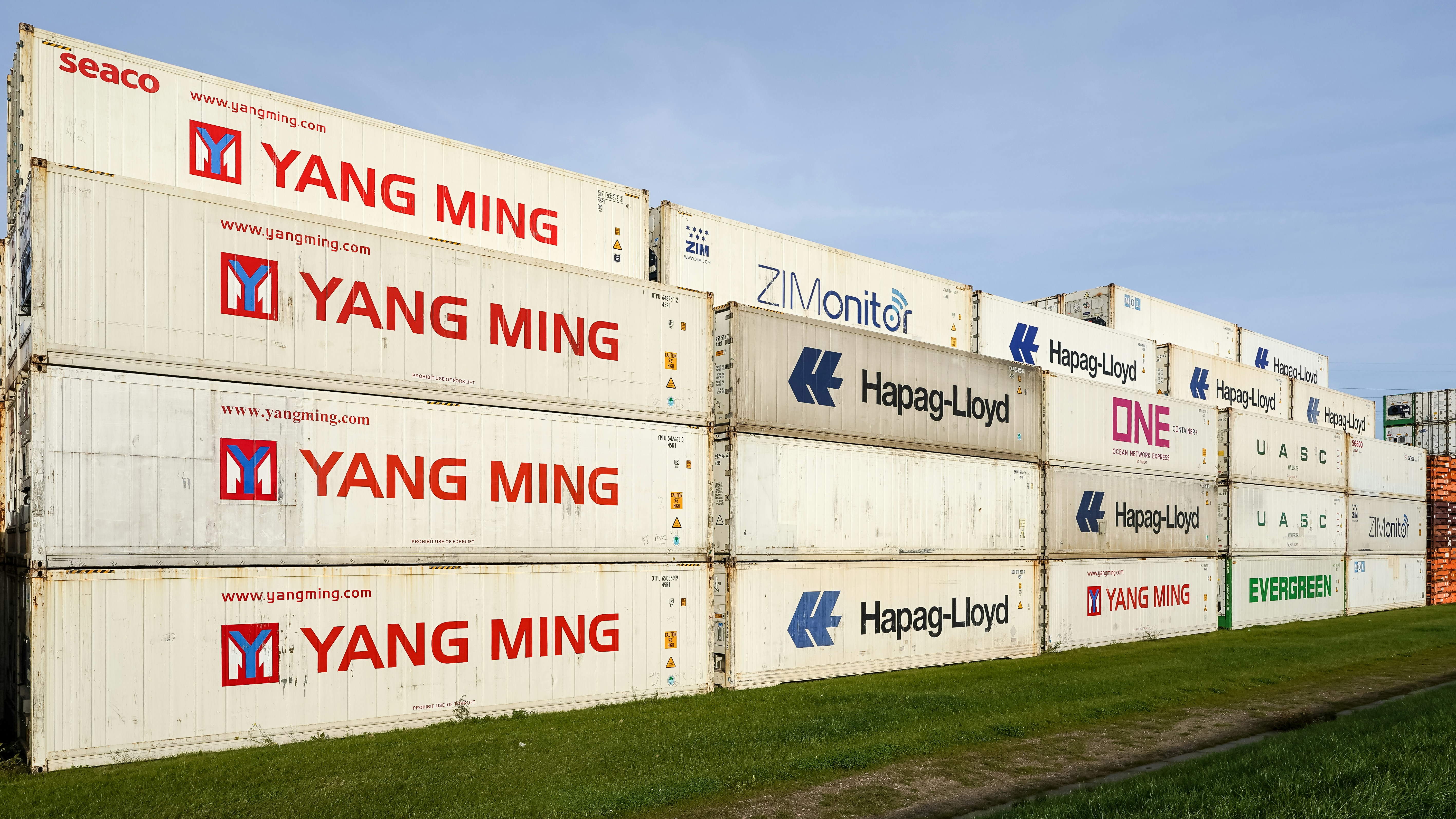 Stacked shipping containers with various company logos on a grassy field under a clear sky