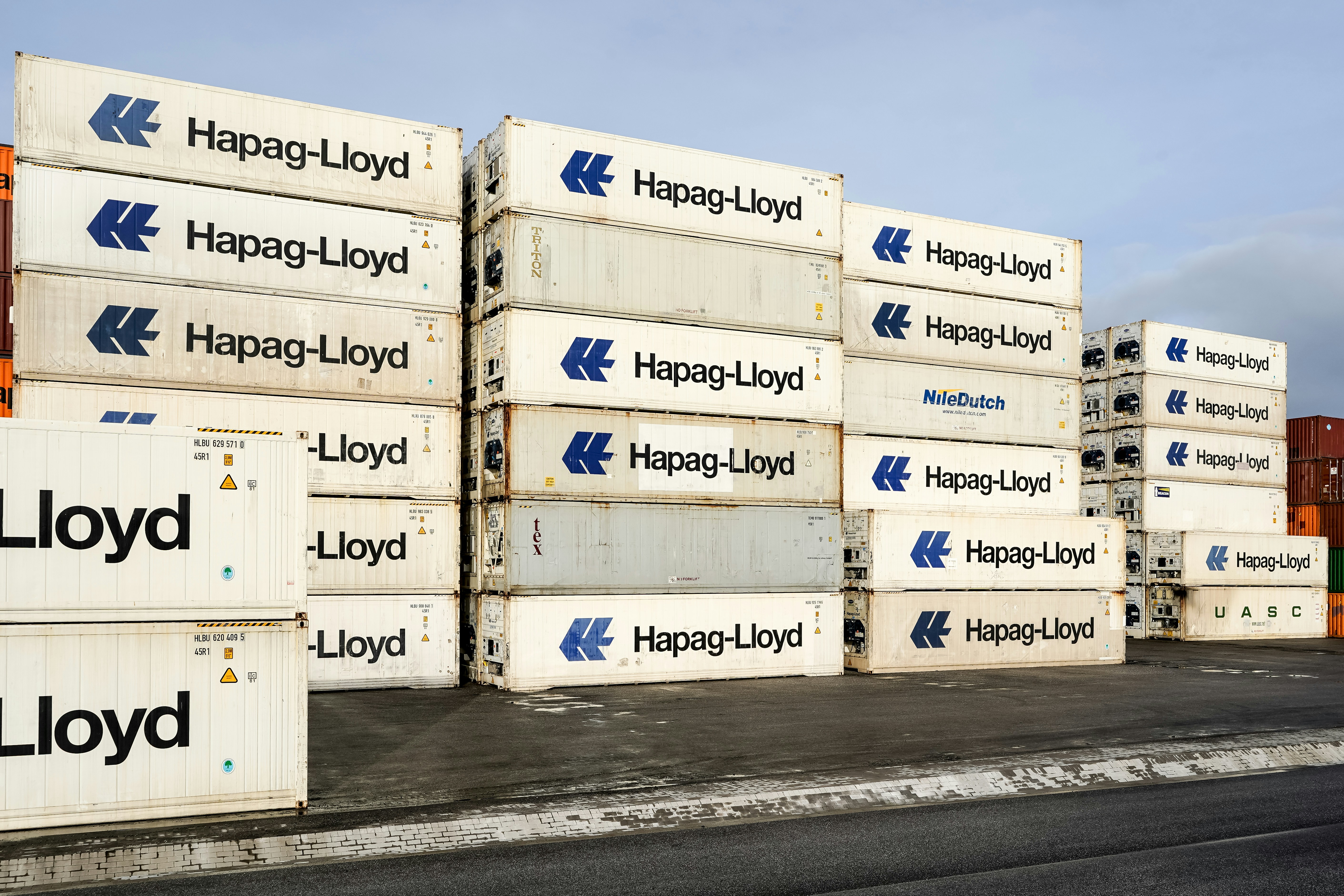 Stacked white shipping containers with Hapag-Lloyd branding in a paved industrial yard