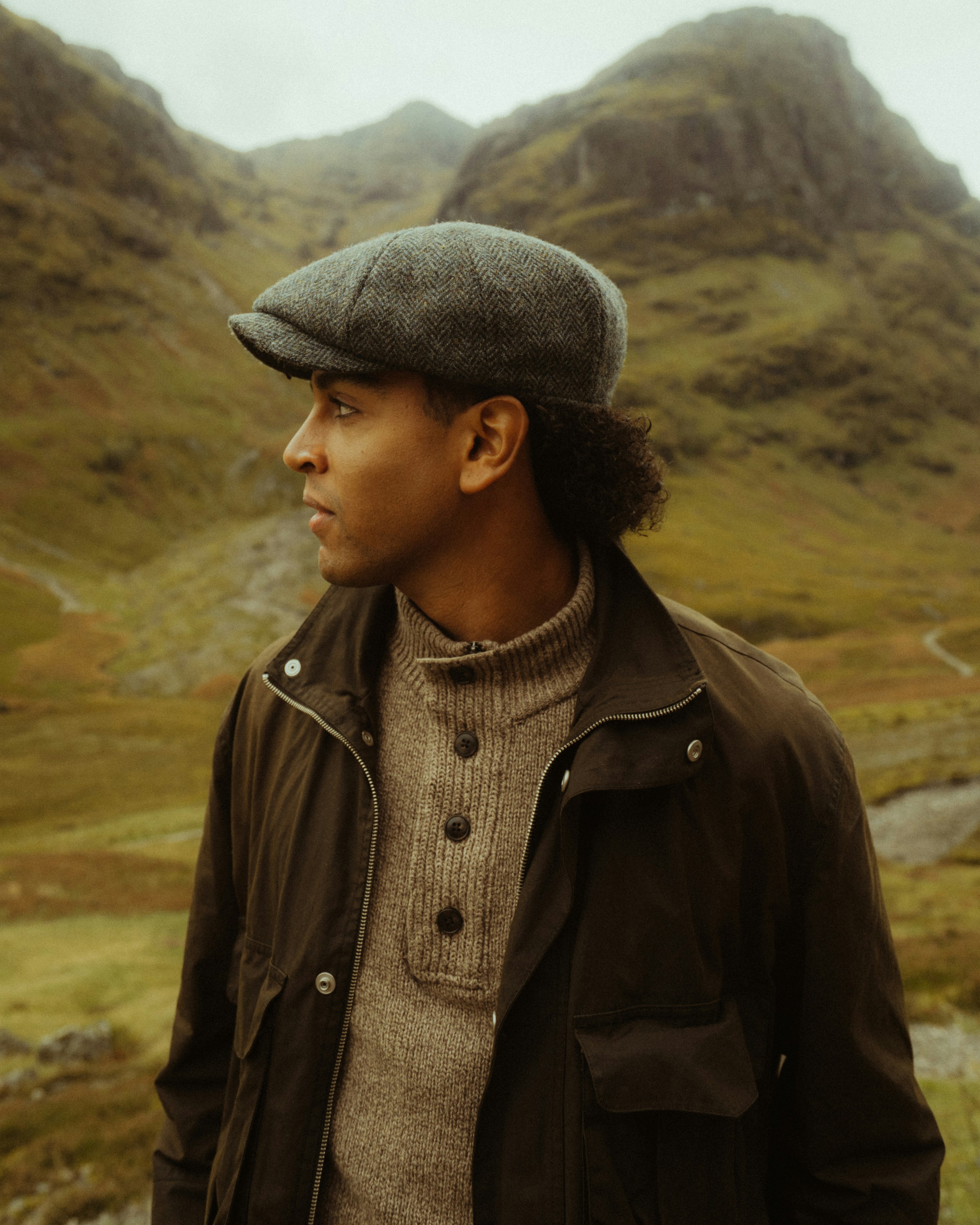 A man in a tweed flat cap and brown jacket against a misty mountain landscape