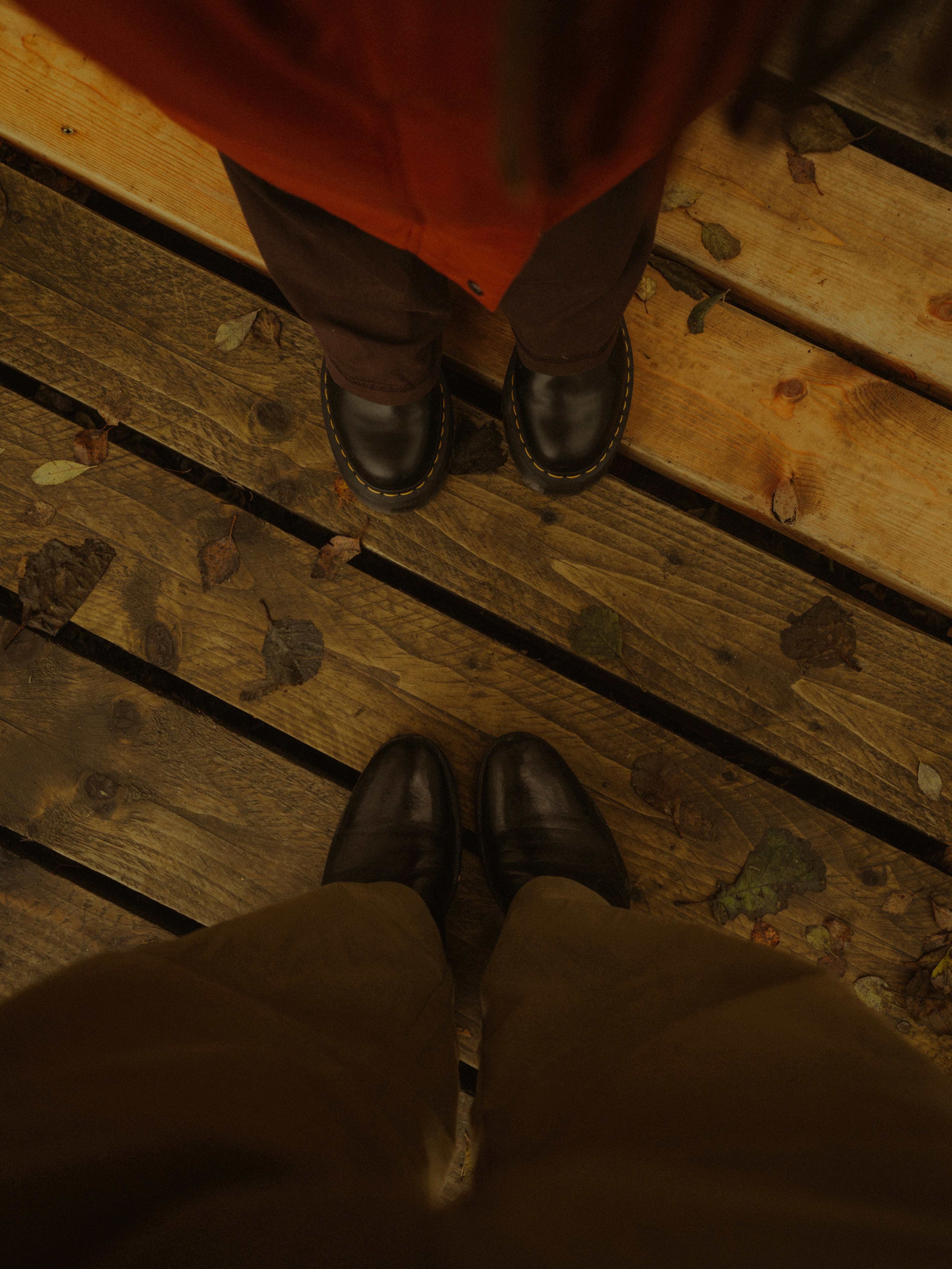 Two people in dark trousers and leather boots standing on a wooden boardwalk with fallen leaves