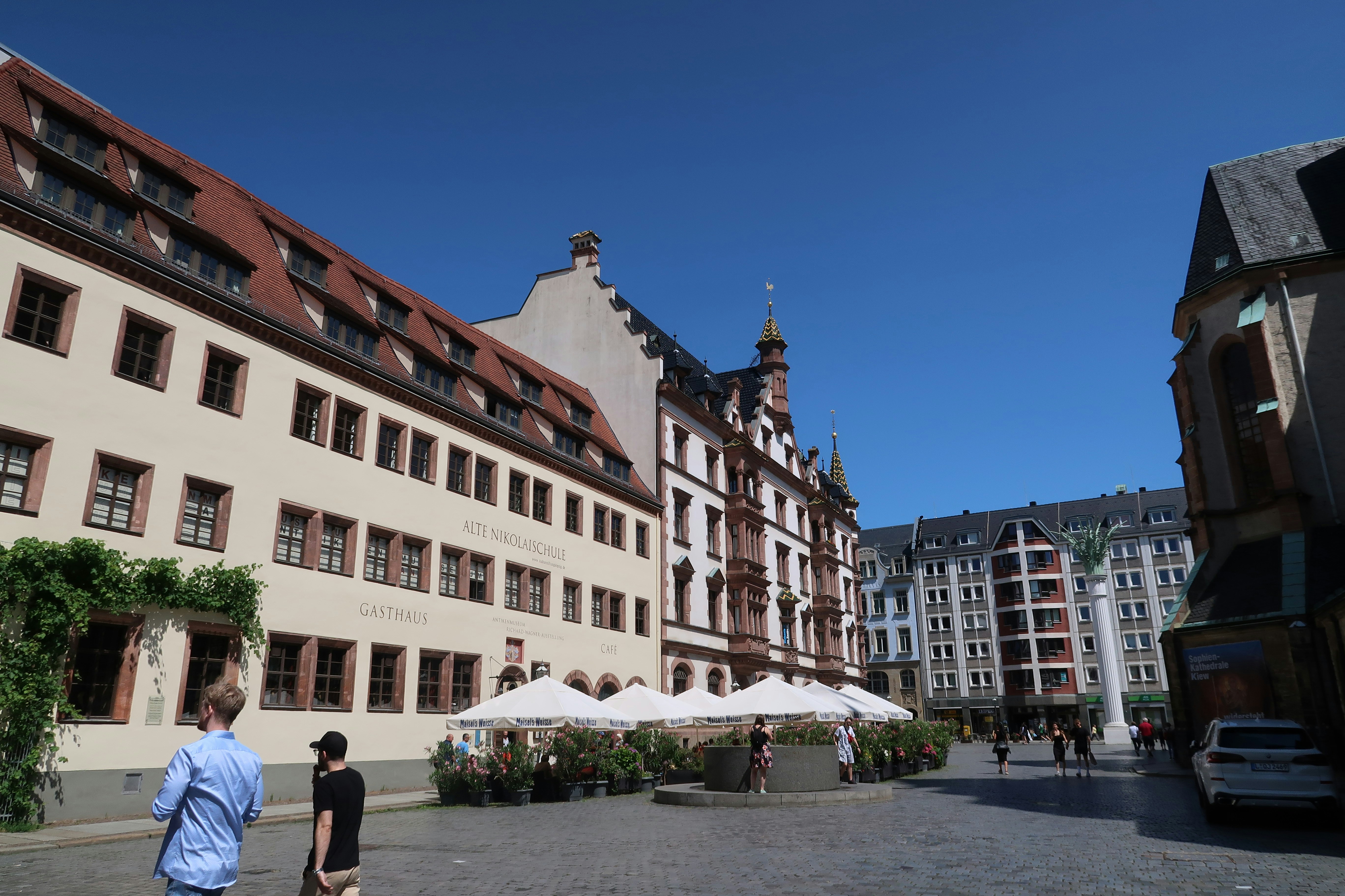Solrig plads i Leipzig med historiske bygninger, gående mennesker og udendørs cafeborde