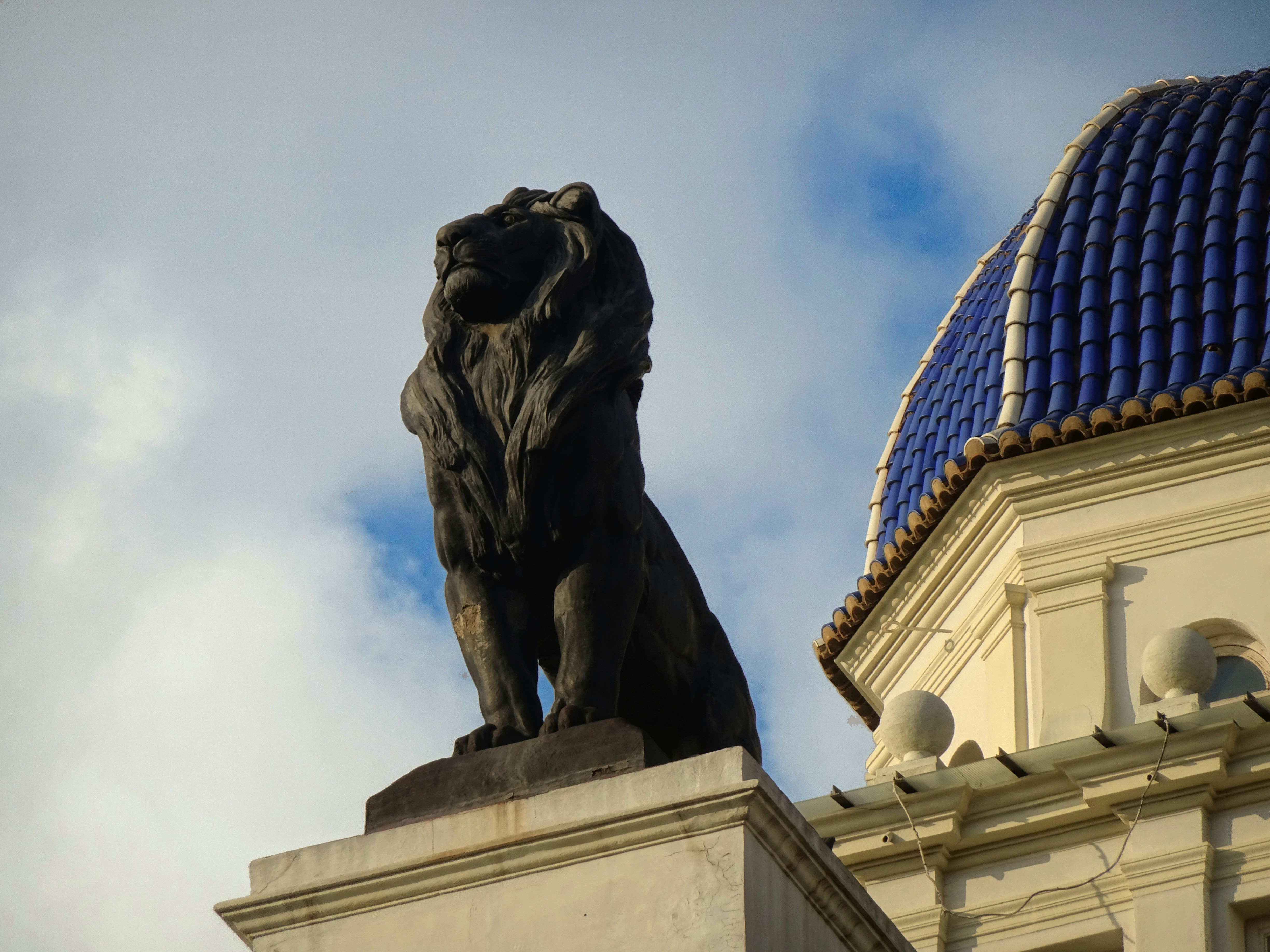 Fotomural A dark bronze lion statue on a stone pedestal beside a building with a blue tiled dome