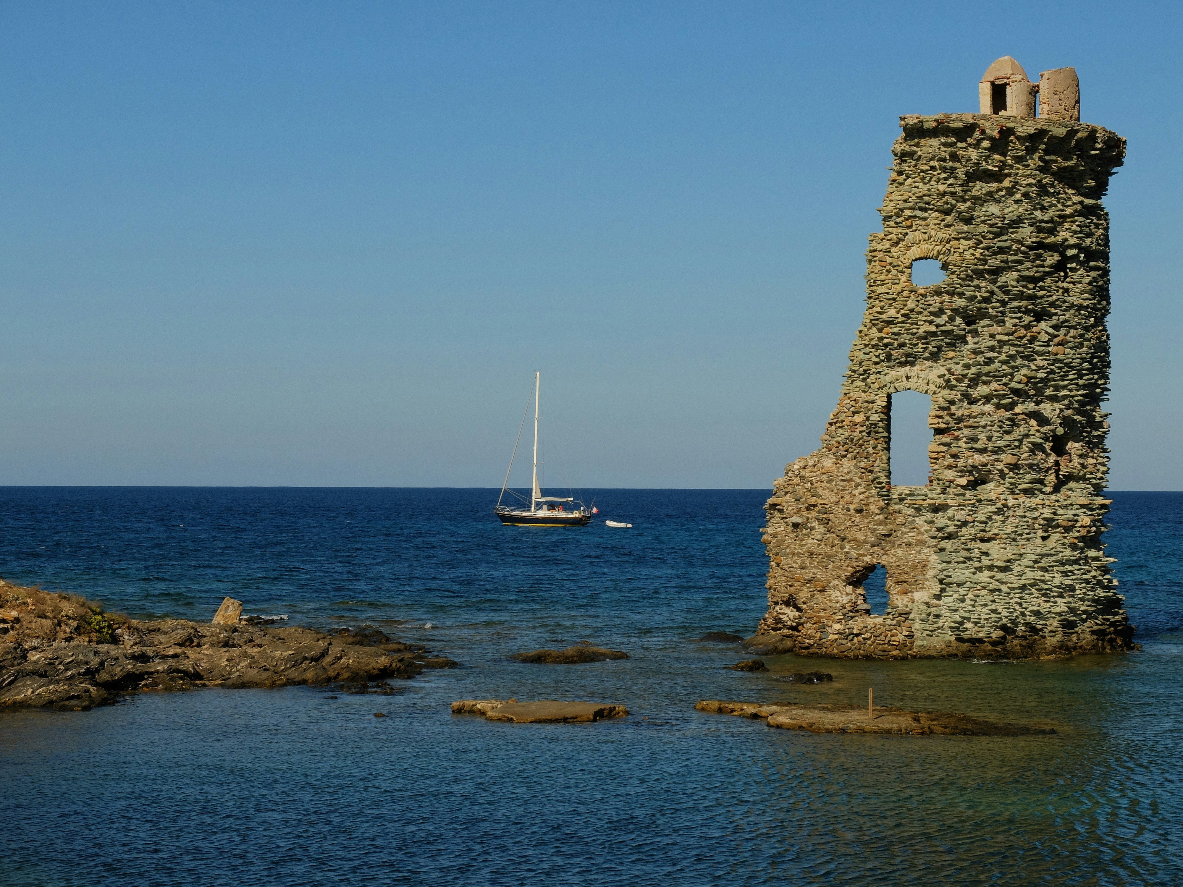 A sailboat on the blue sea near a ruined stone tower on a rocky coast