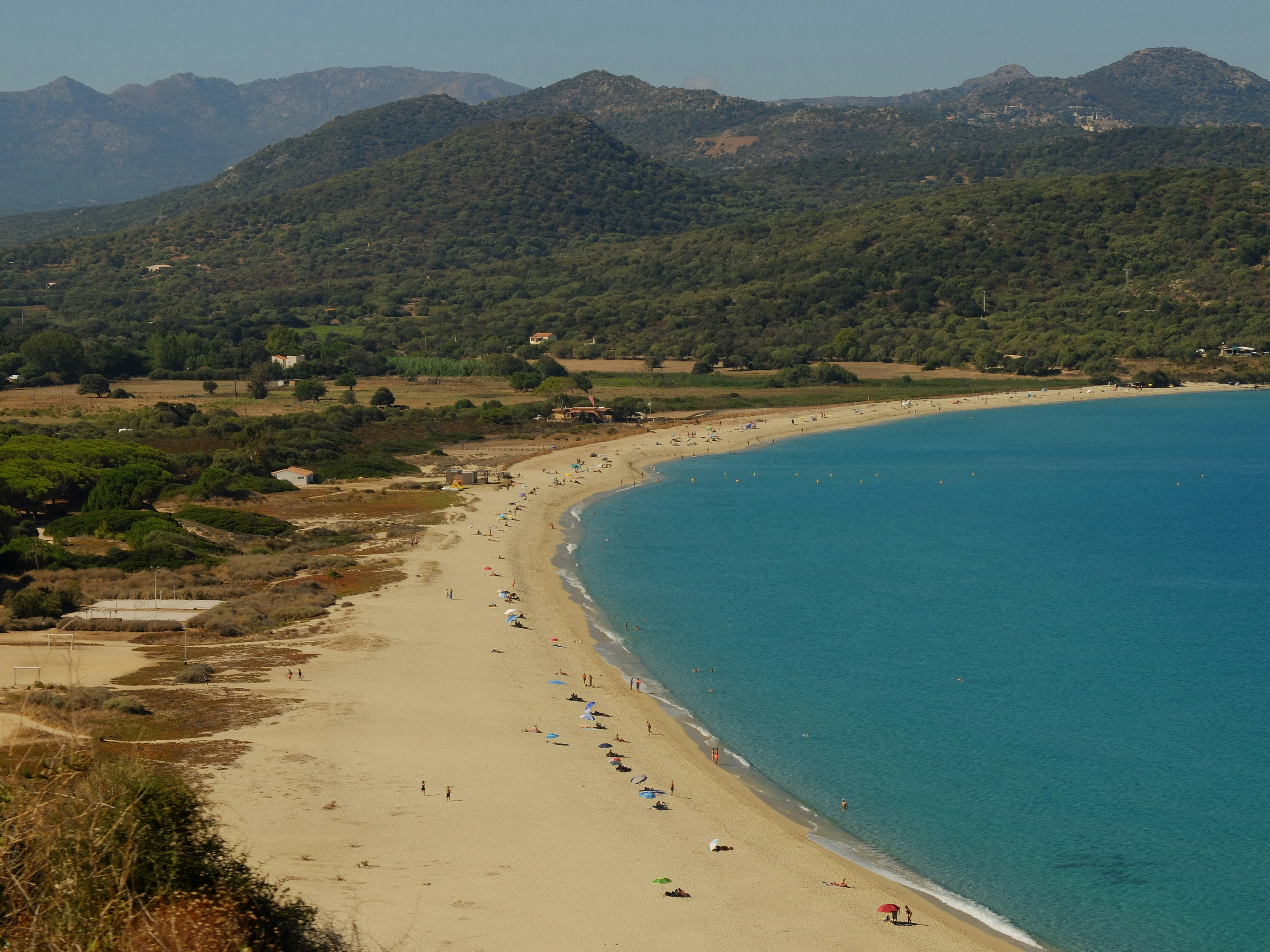 A sandy beach with people and umbrellas along a turquoise bay under green hills