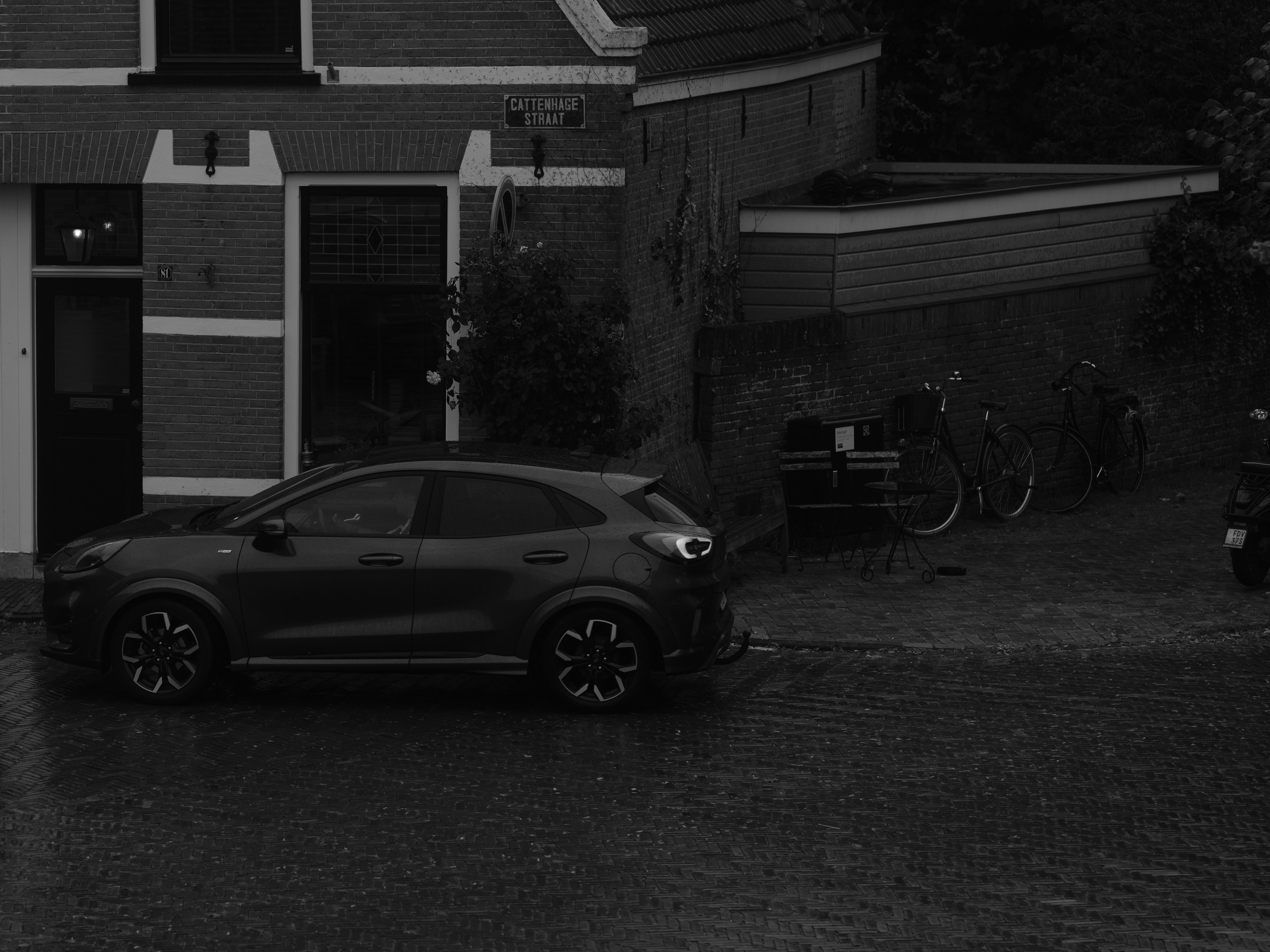 Sebuah mobil gelap terparkir di jalan berbatu di samping bangunan bata dengan warna hitam putih