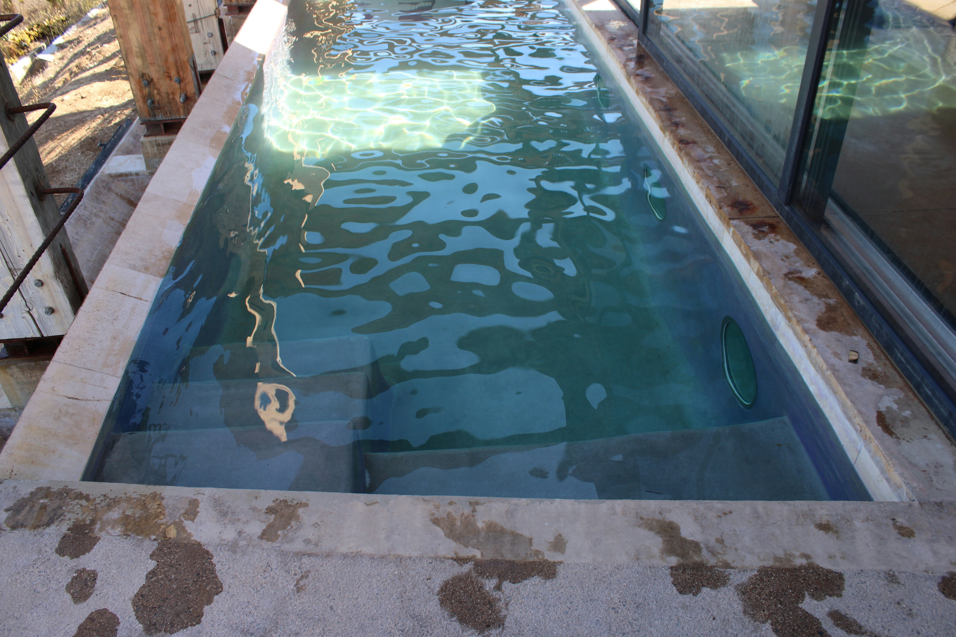 A rectangular swimming pool with clear water and submerged steps beside a glass wall
