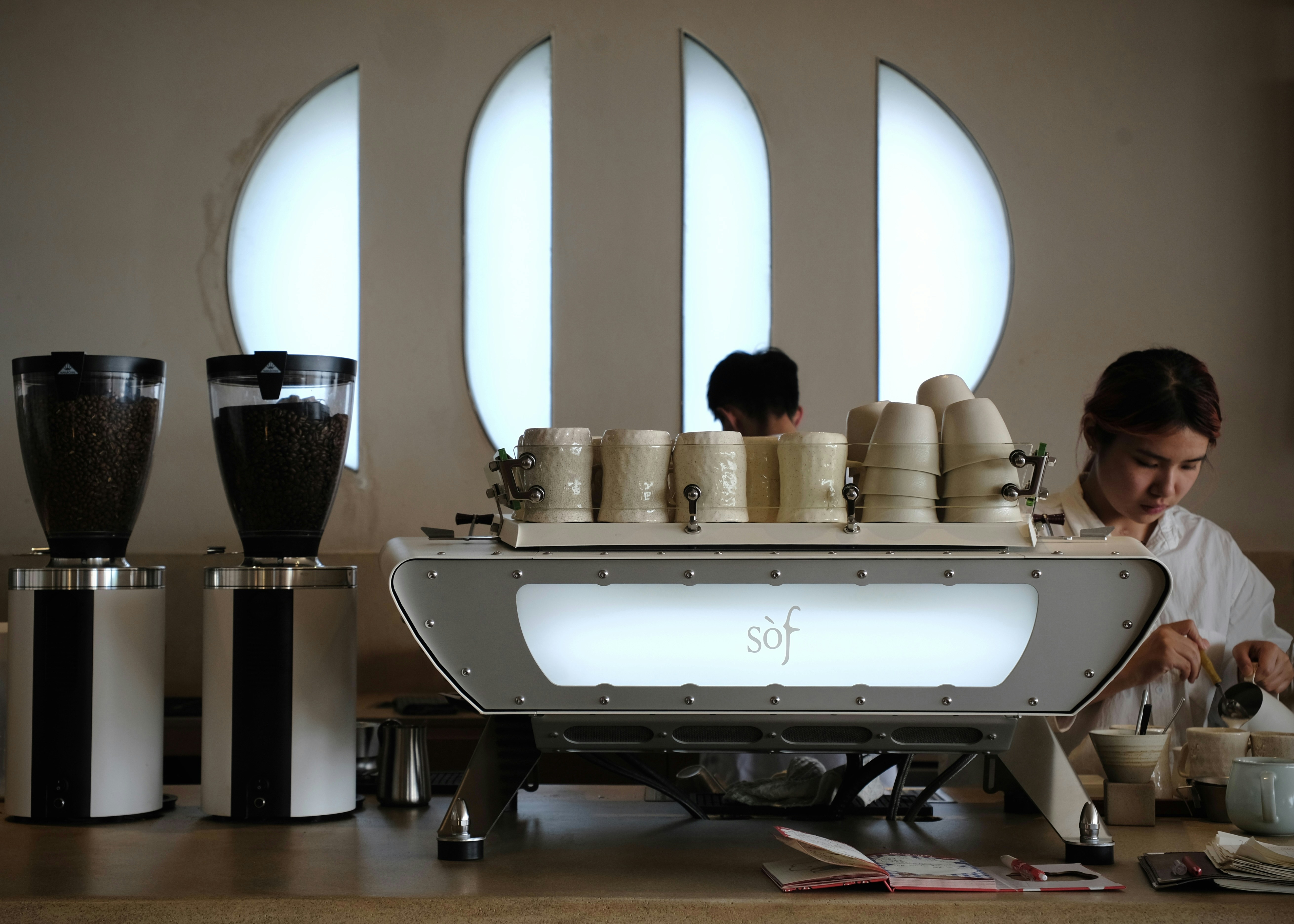 A barista preparing coffee behind a white espresso machine in a cafe with arched windows
