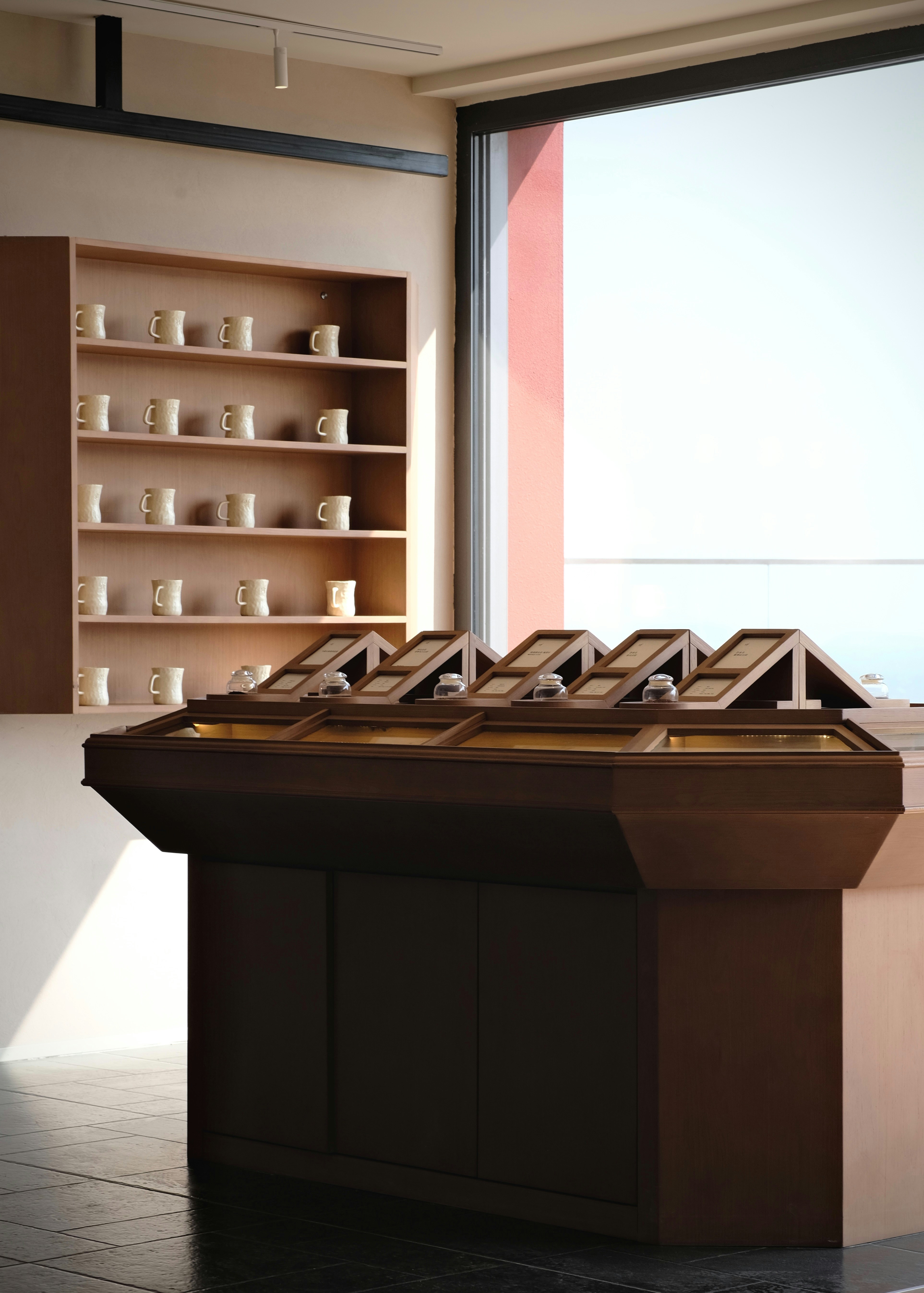 A wooden display counter with glass cases beside shelves of ceramic mugs in a sunlit room
