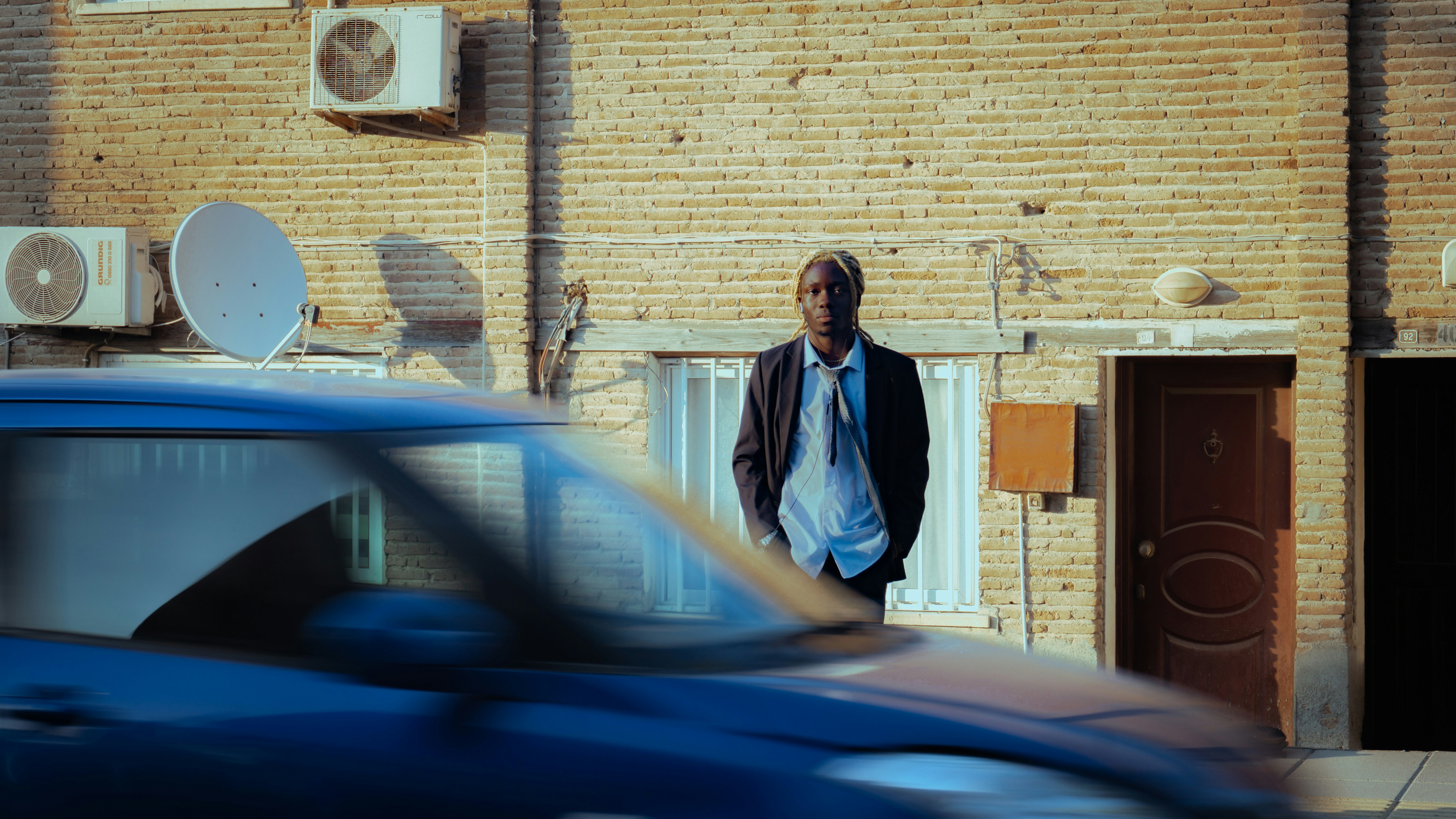 A man in a dark suit standing before a brick wall as a blue car blurs past