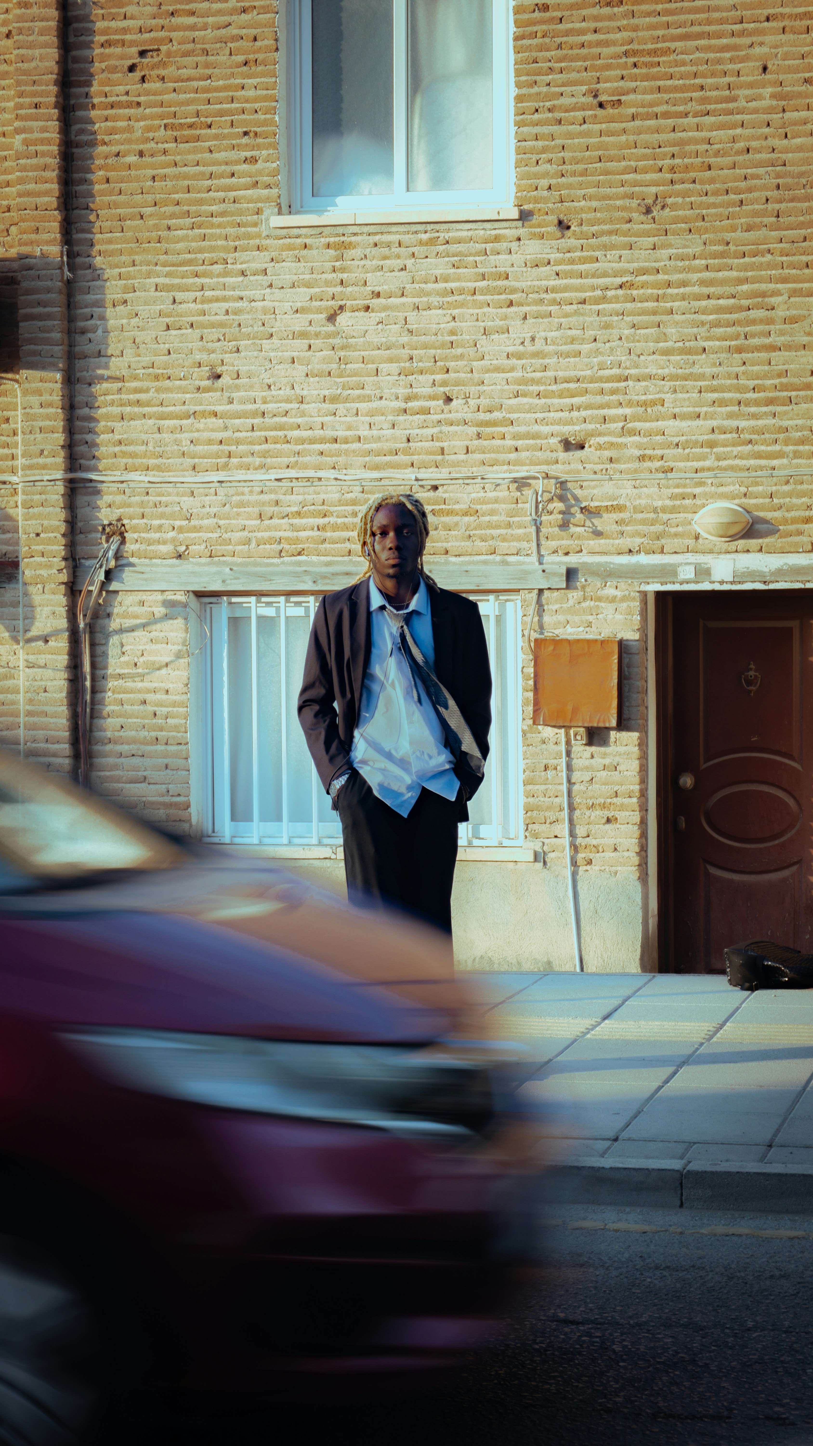 A person in a suit and tie standing before a brick building with a blurred car passing