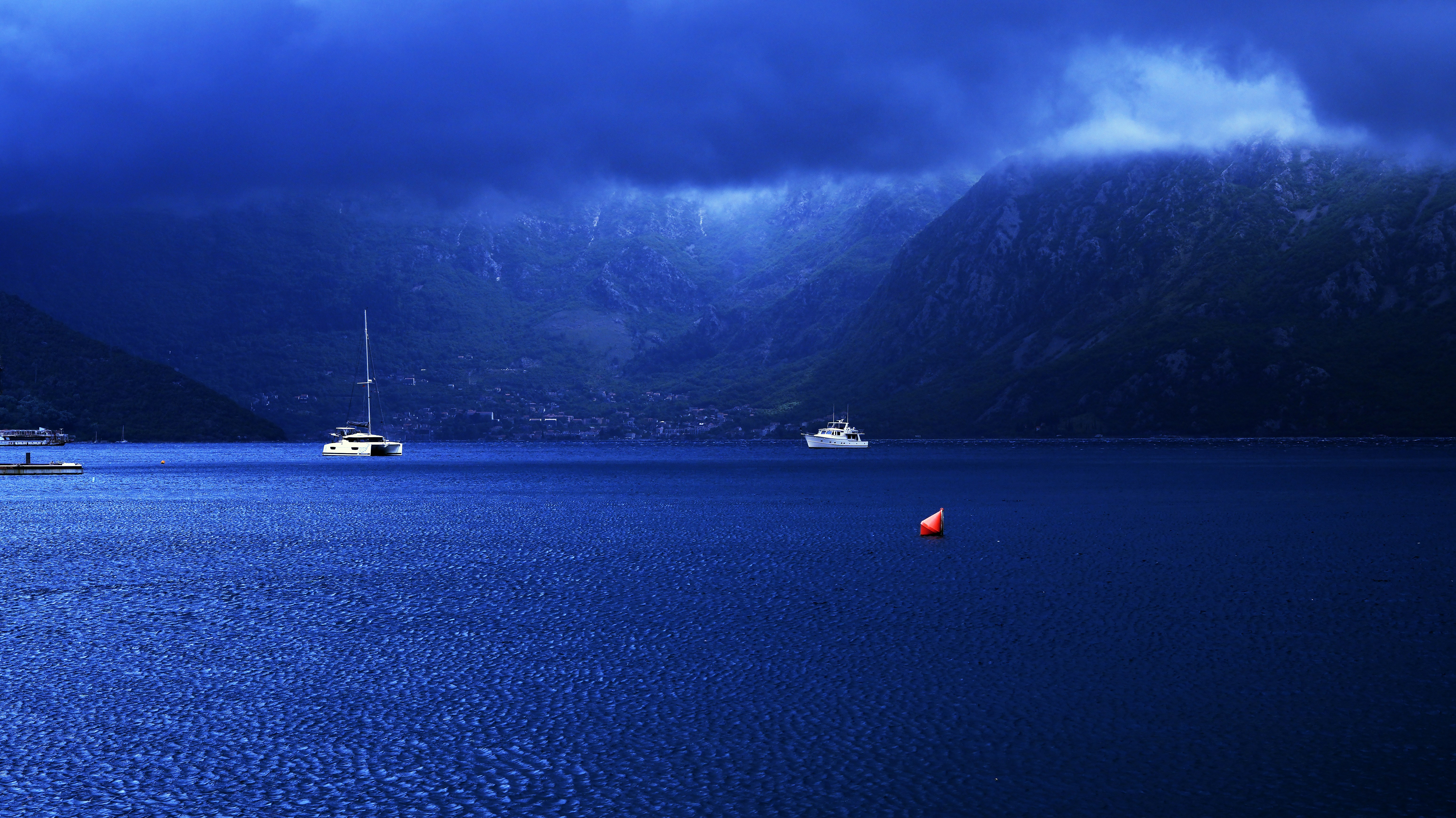 Boats on a deep blue bay beneath dark mountains shrouded in heavy clouds
