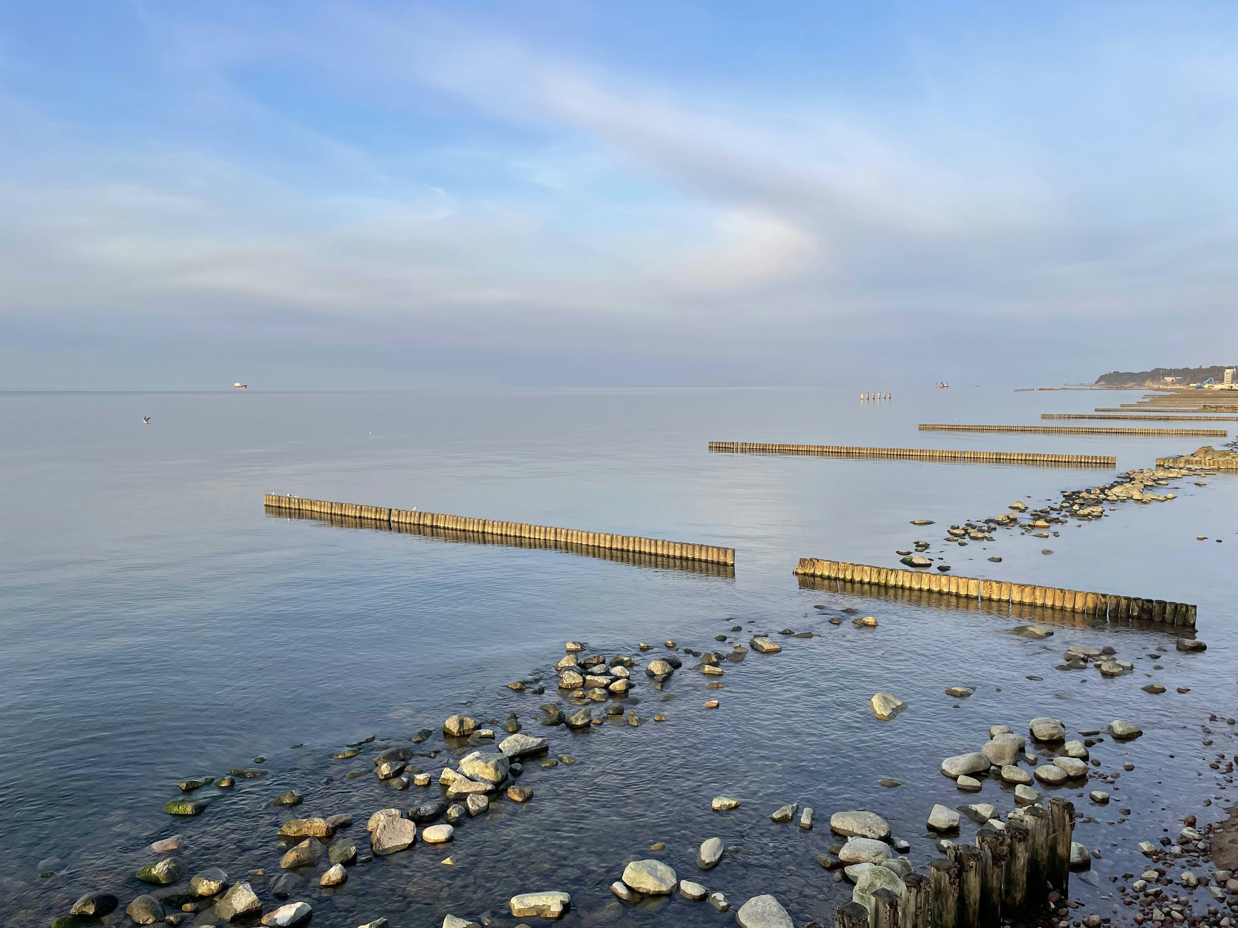 Wooden groynes extending into calm sea water with a rocky shoreline under a soft blue sky