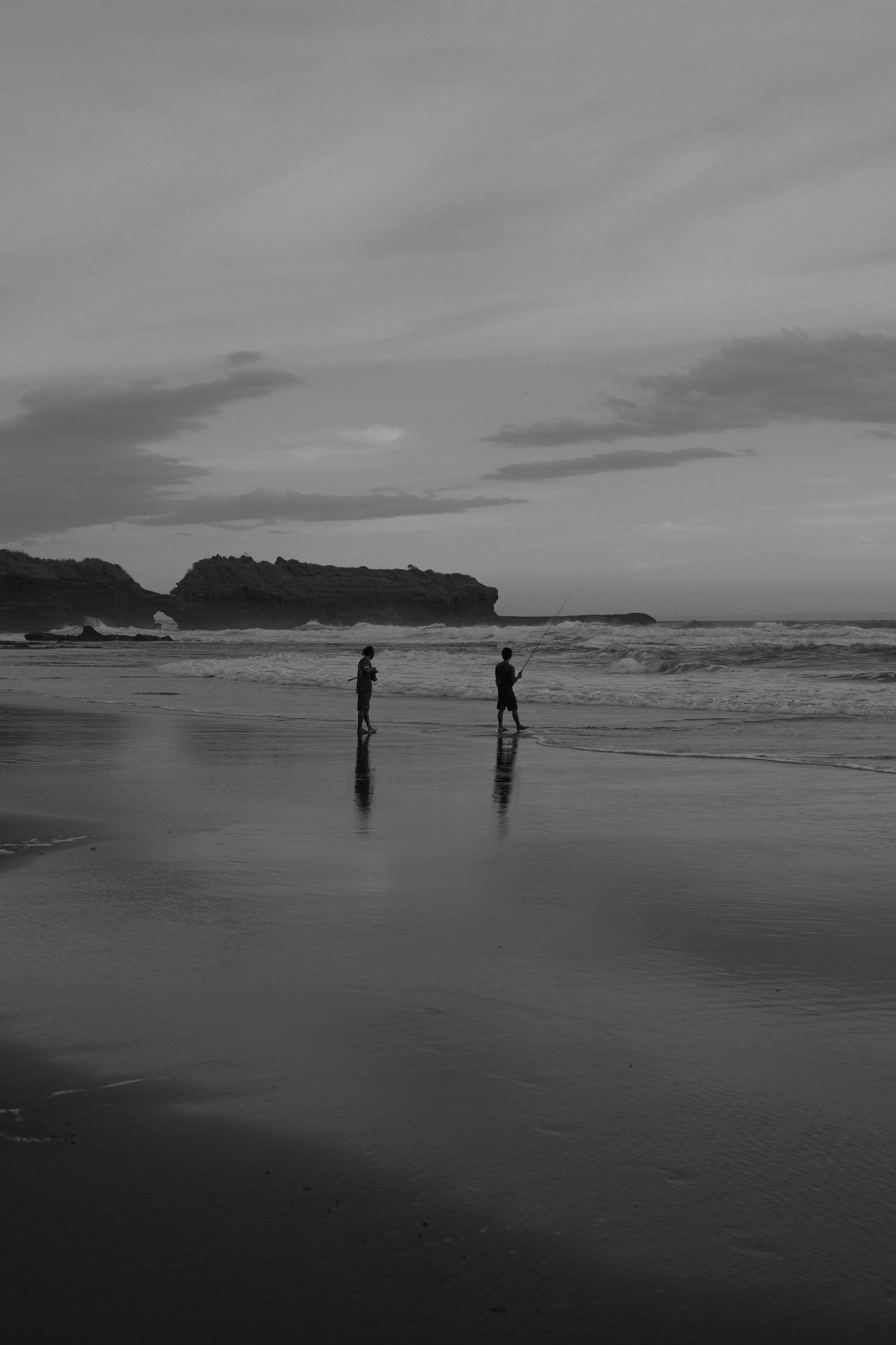Two people fishing in the surf on a beach under a cloudy sky, black and white