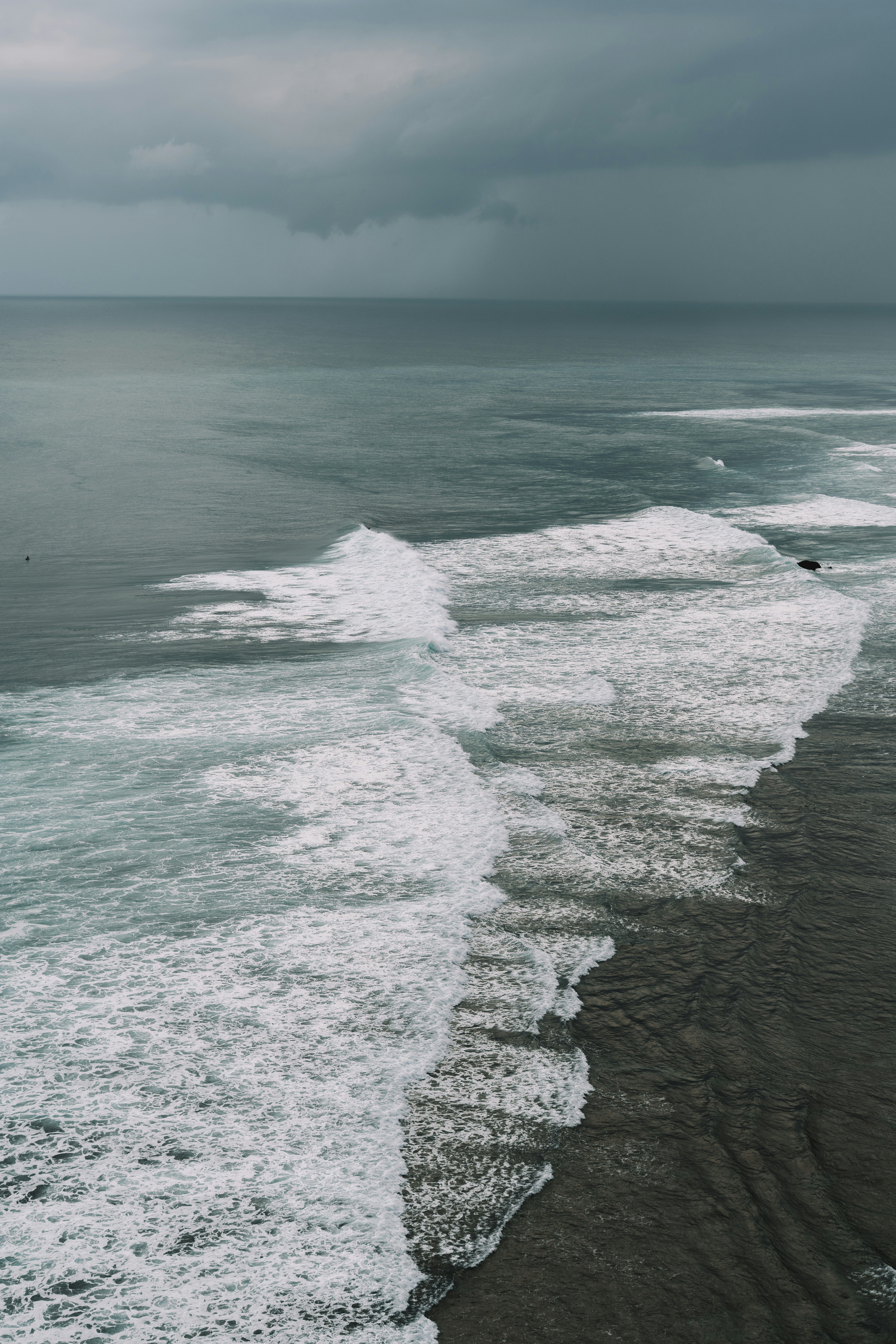 Aerial view of white waves breaking on a dark rocky shoreline under a stormy grey sky