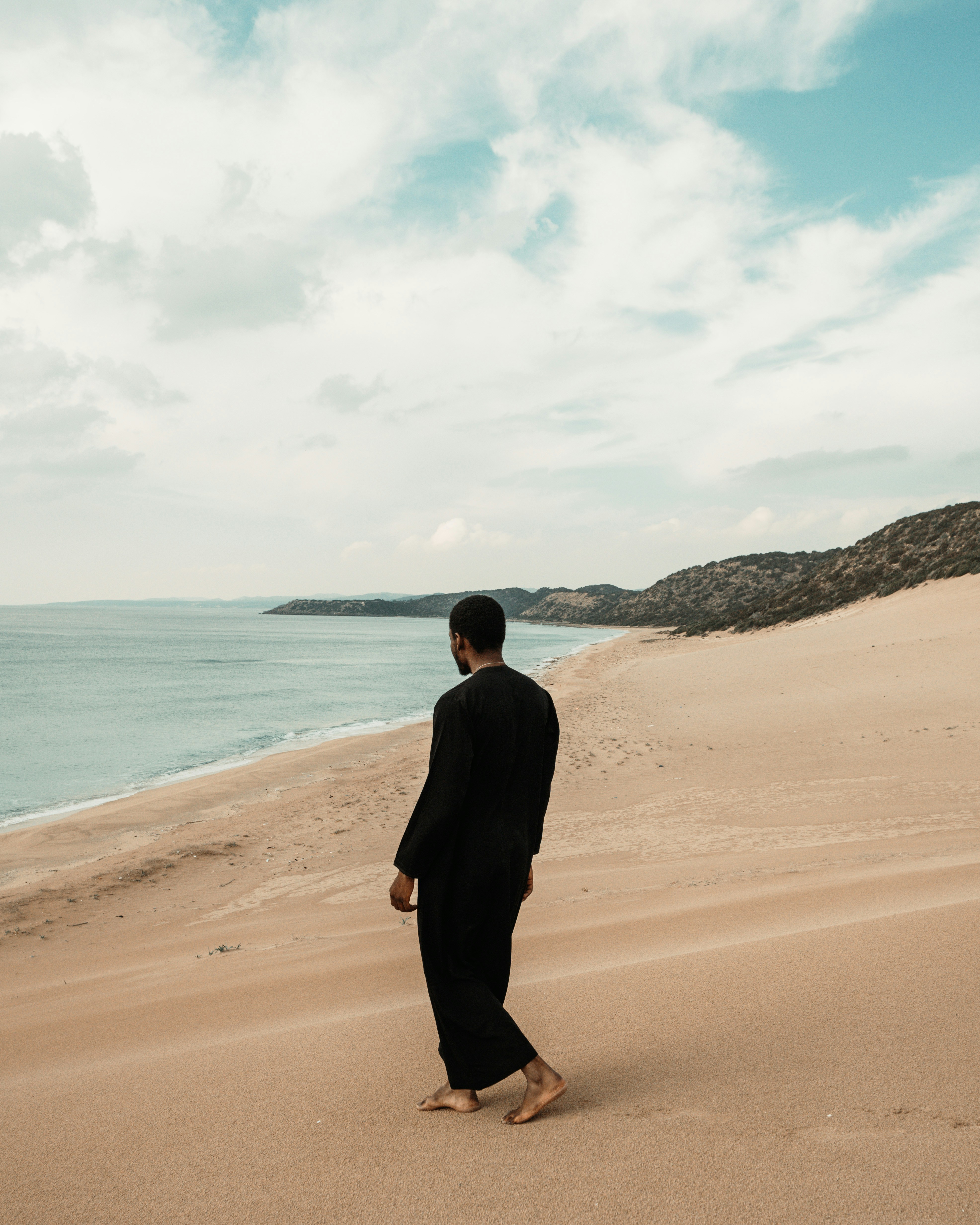 A person in a black robe walking barefoot on a sandy beach by the sea