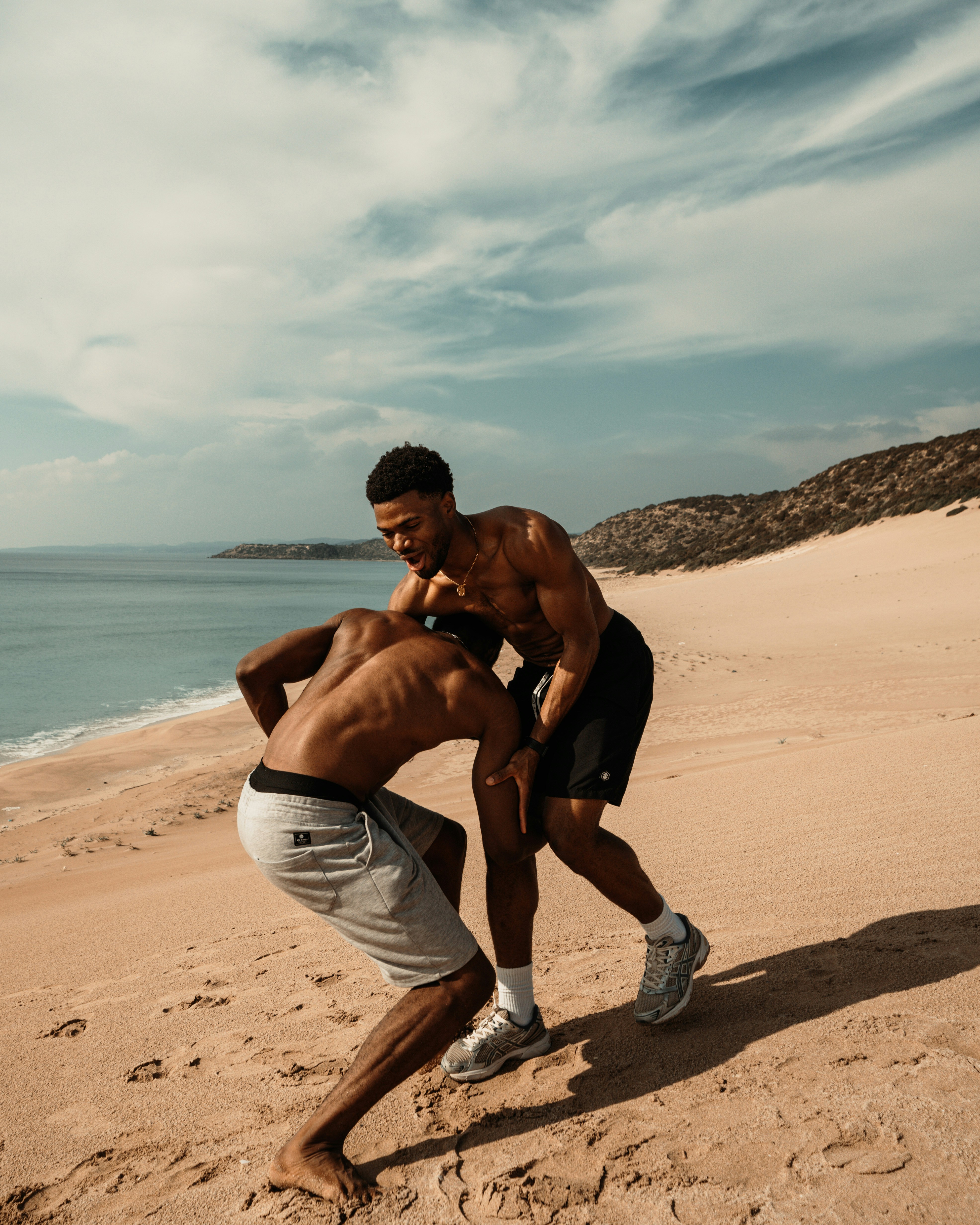 Deux lutteurs torse nu s'affrontent sur une plage de sable sous un ciel clair, une scène proche de la lutte de plage