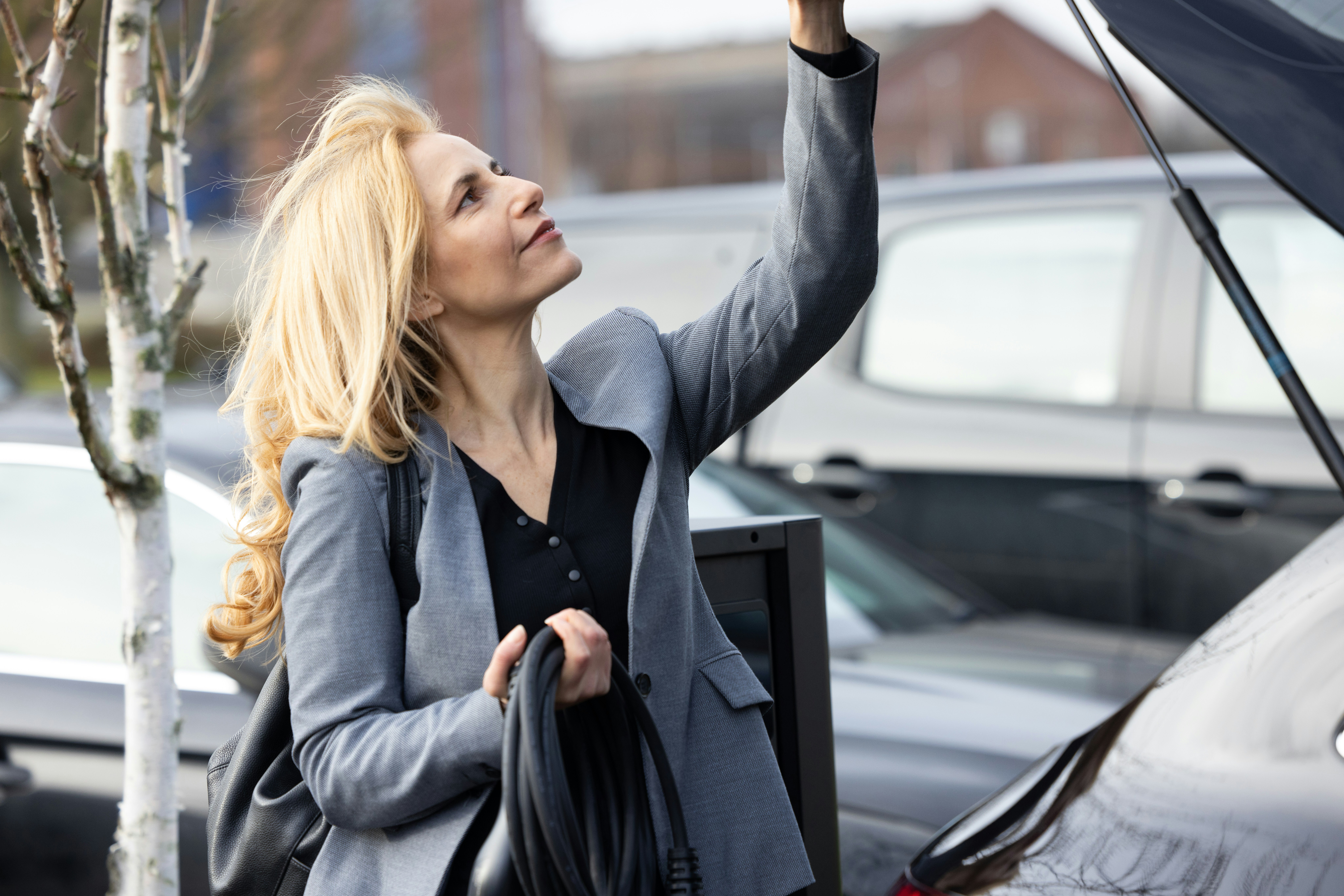 A woman in a grey blazer holding a charging cable at an electric vehicle station