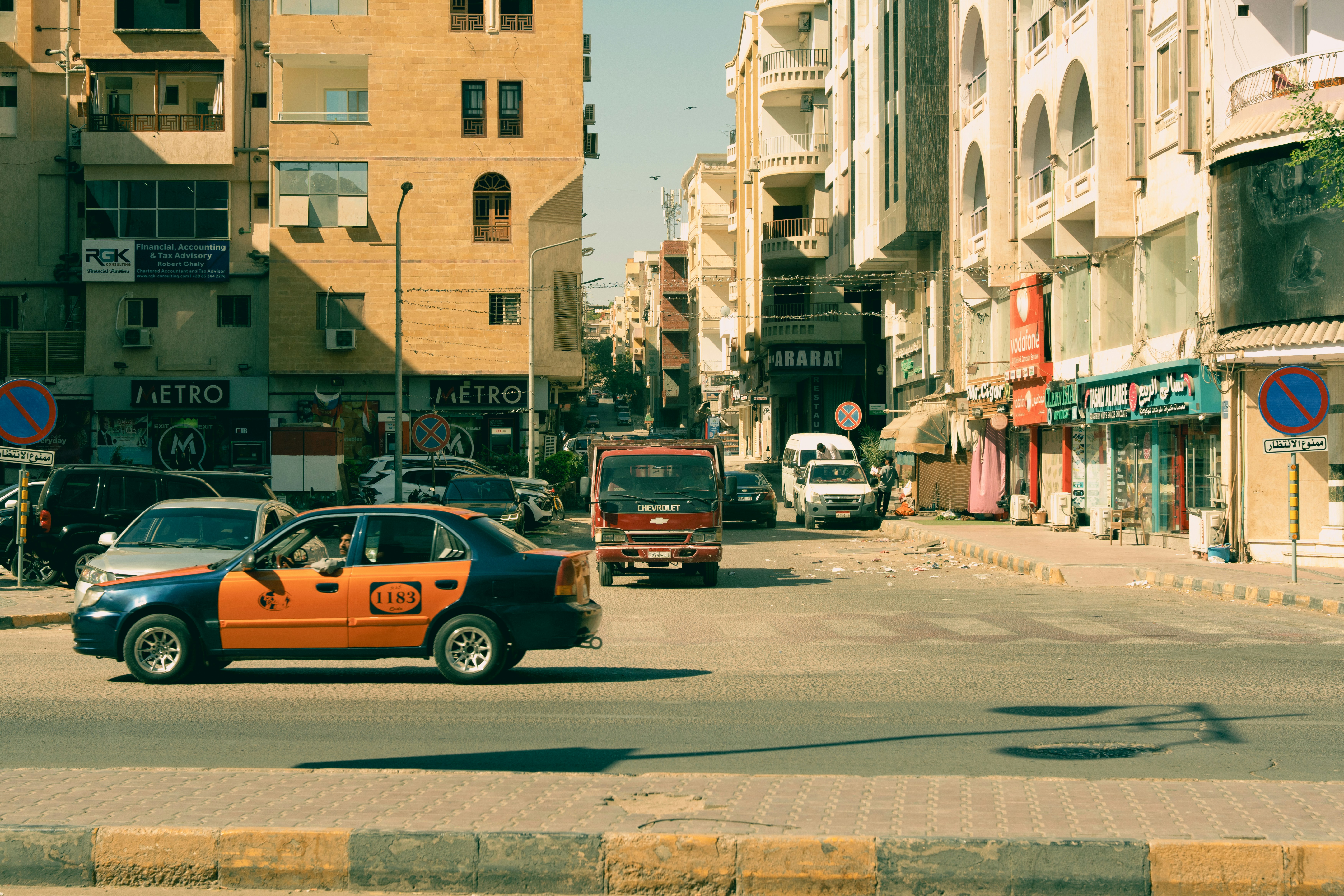 A blue and orange taxi driving down a busy city street lined with shops and apartment buildings