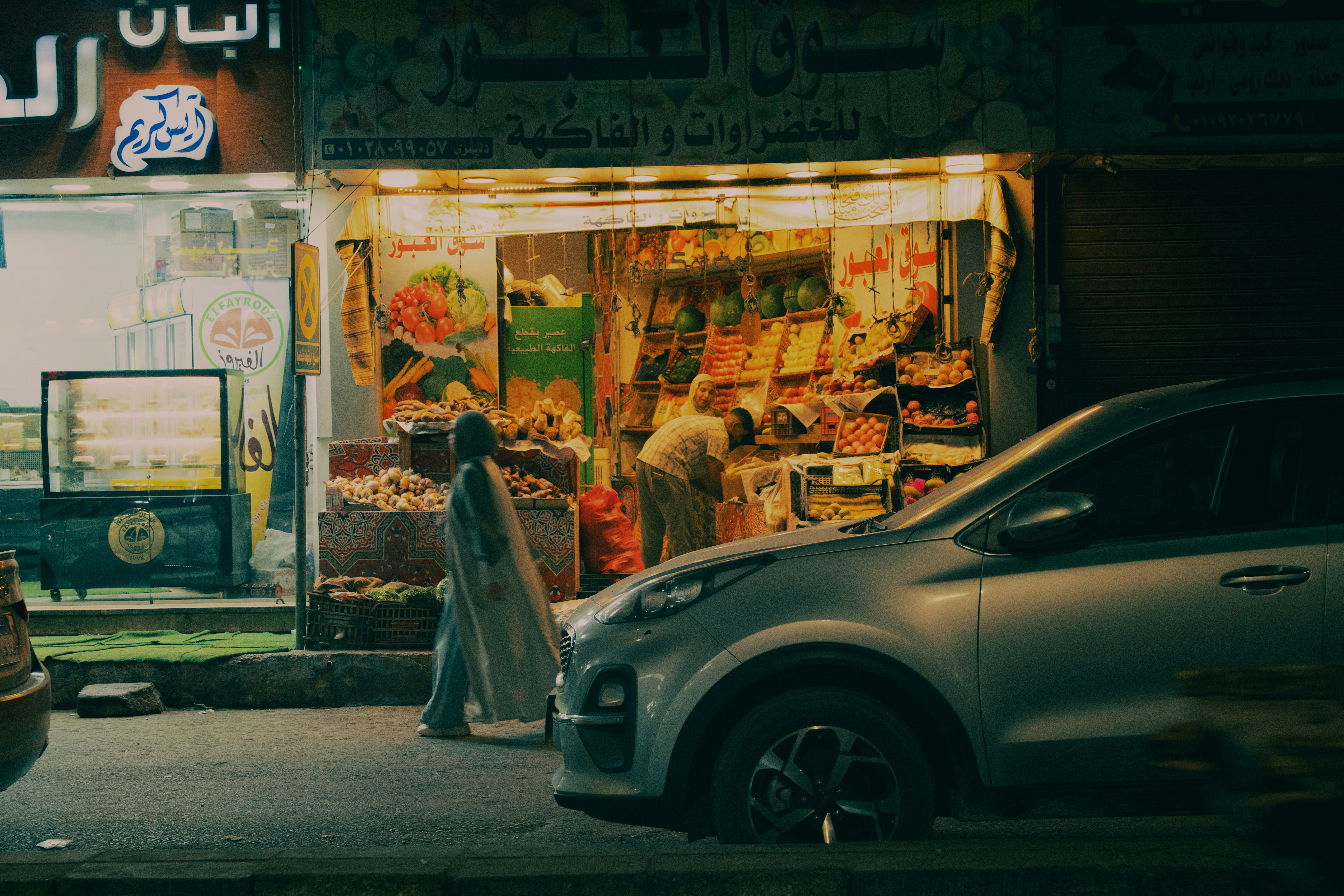 A person in a long coat walking past a brightly lit fruit and vegetable shop at night