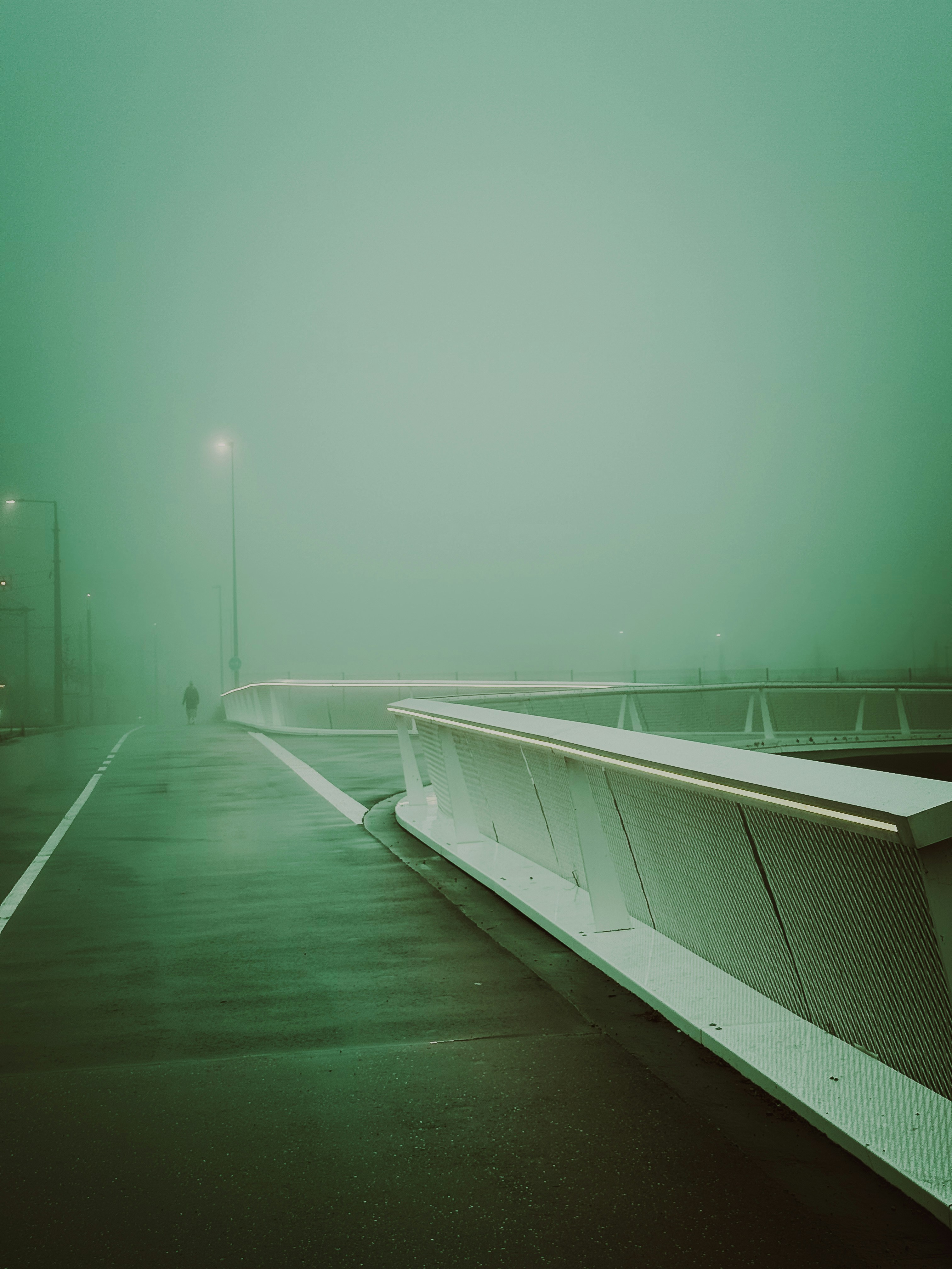 A person walking on a wet path beside a white bridge in thick green fog