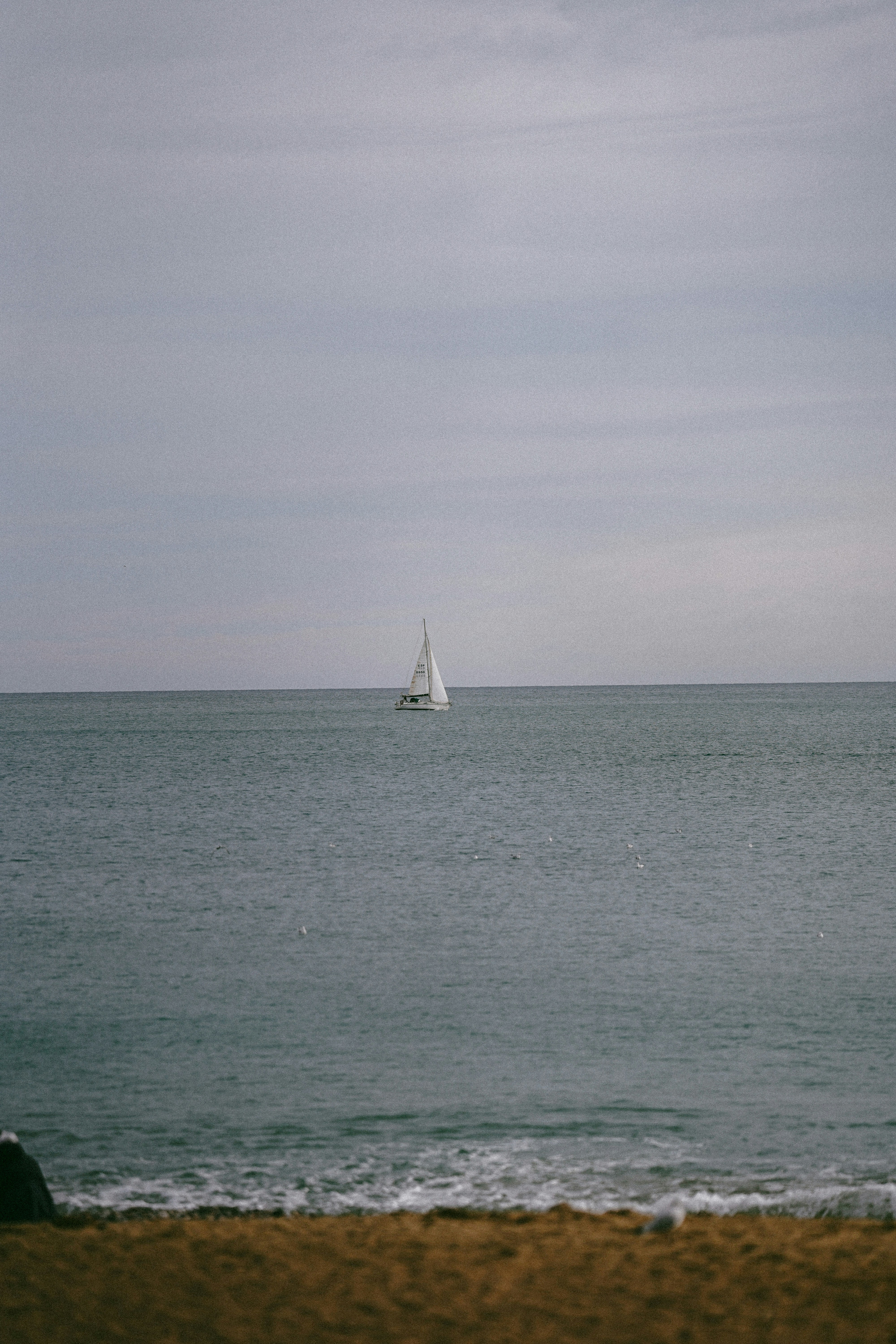 A lone sailboat on the calm grey sea under an overcast sky