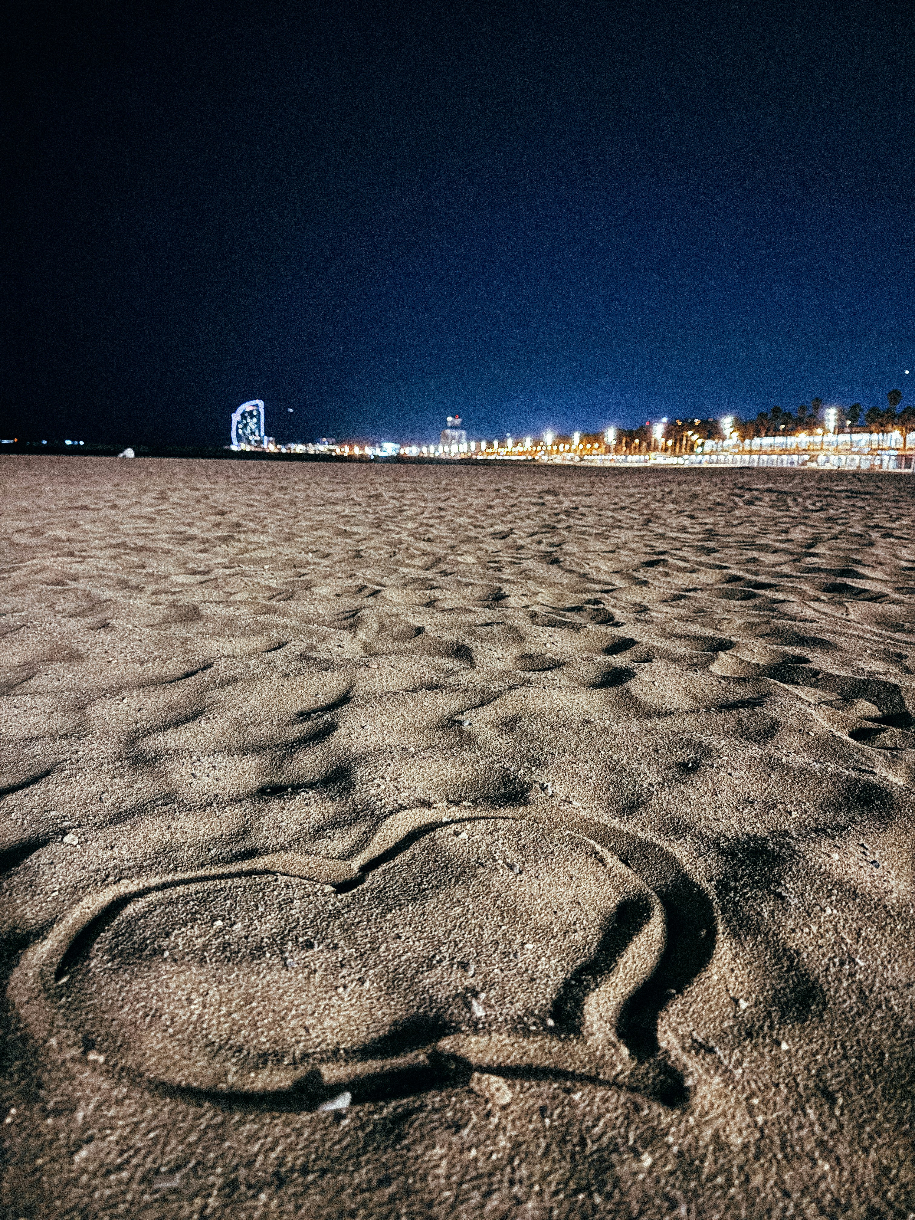 A heart drawn in the sand on a beach at night with city lights in the distance