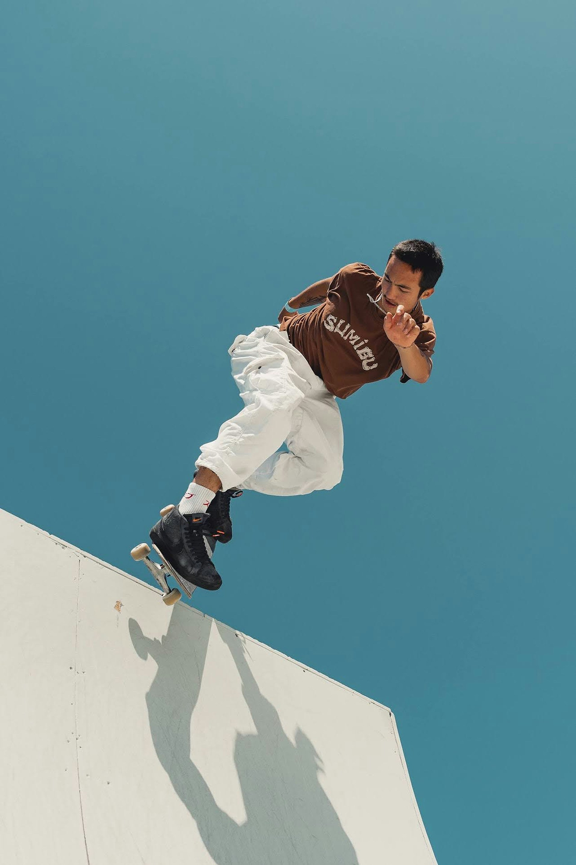 A man in a brown t-shirt and white pants performing a skateboard trick against a clear blue sky