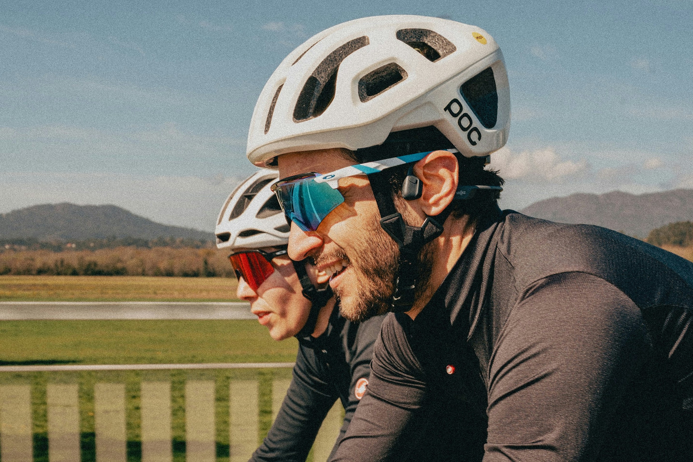 Two cyclists in helmets and sunglasses riding along a road with mountains in the distance