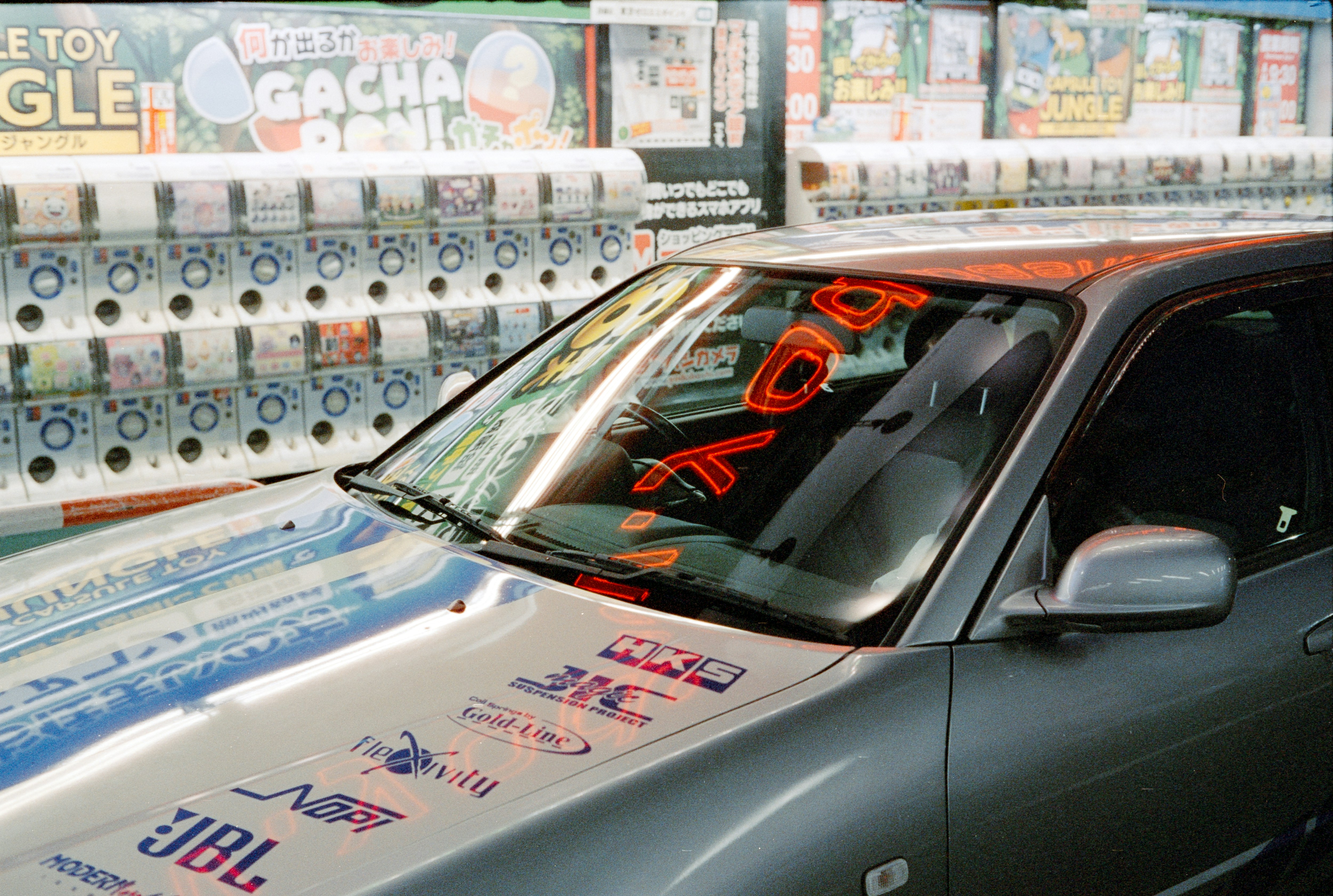 A grey car with racing decals parked in front of a row of capsule toy machines