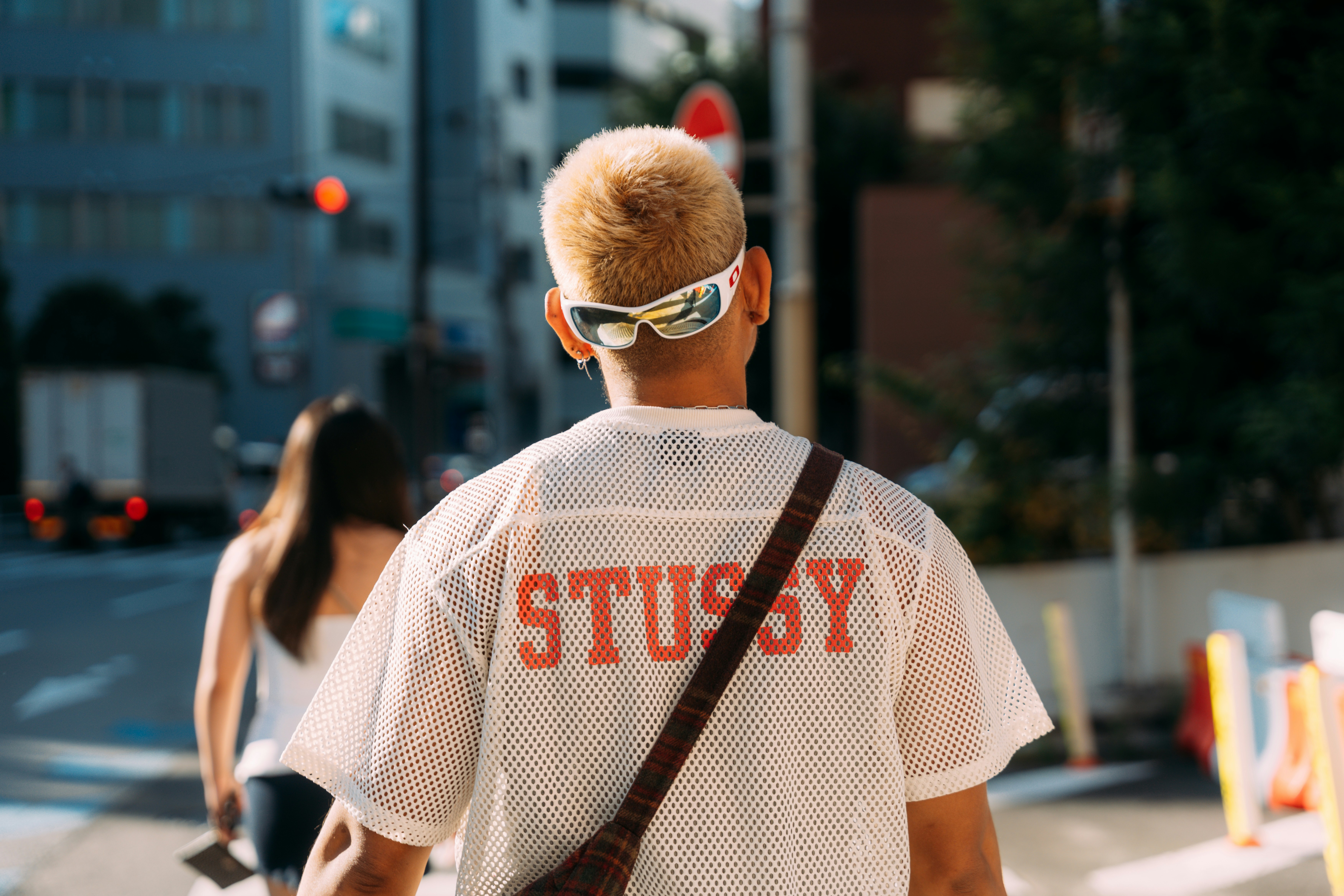 A person with blonde hair wearing white sunglasses and a mesh shirt on a city street