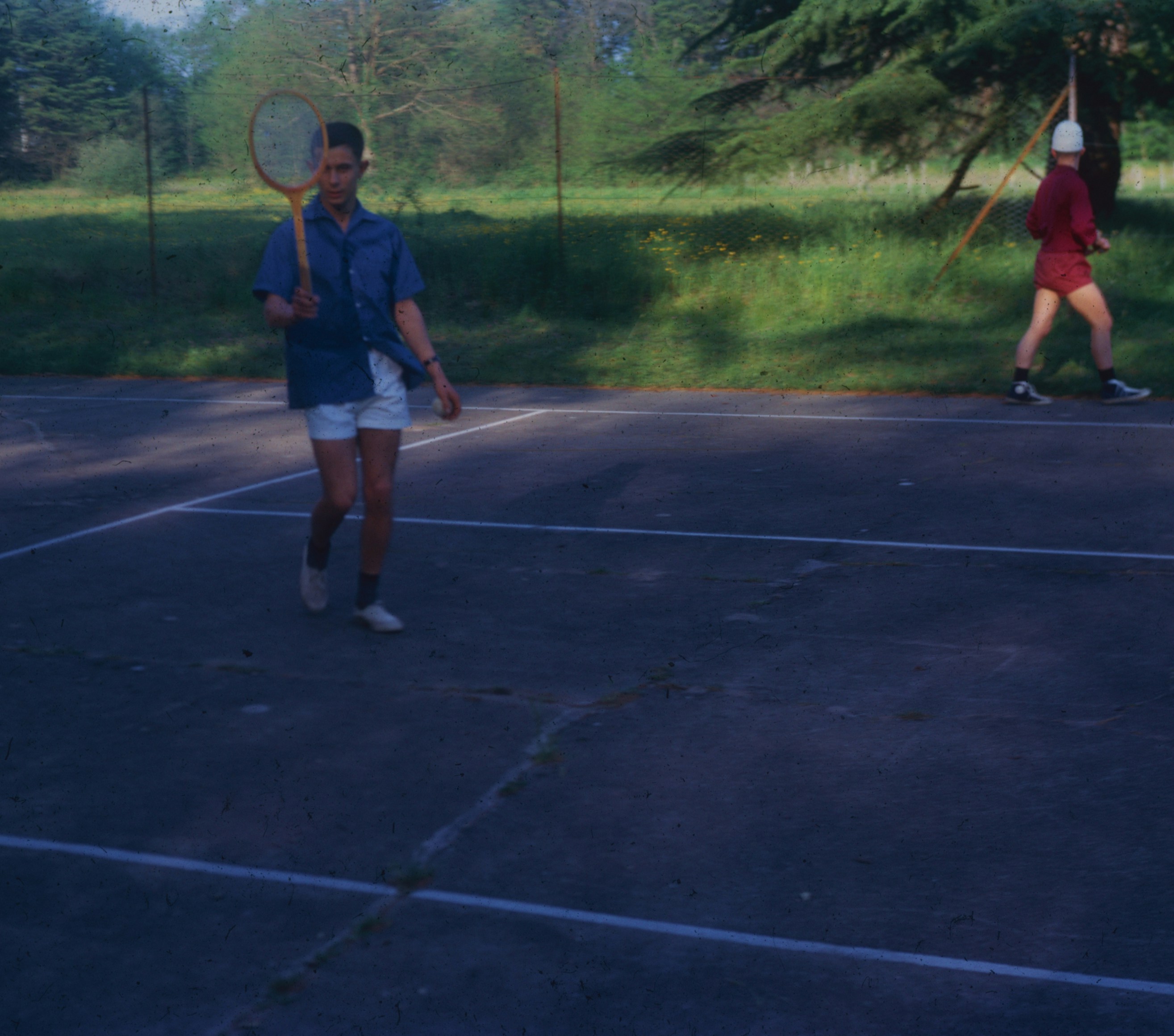 A person holding a badminton racket on a paved court with another person walking nearby