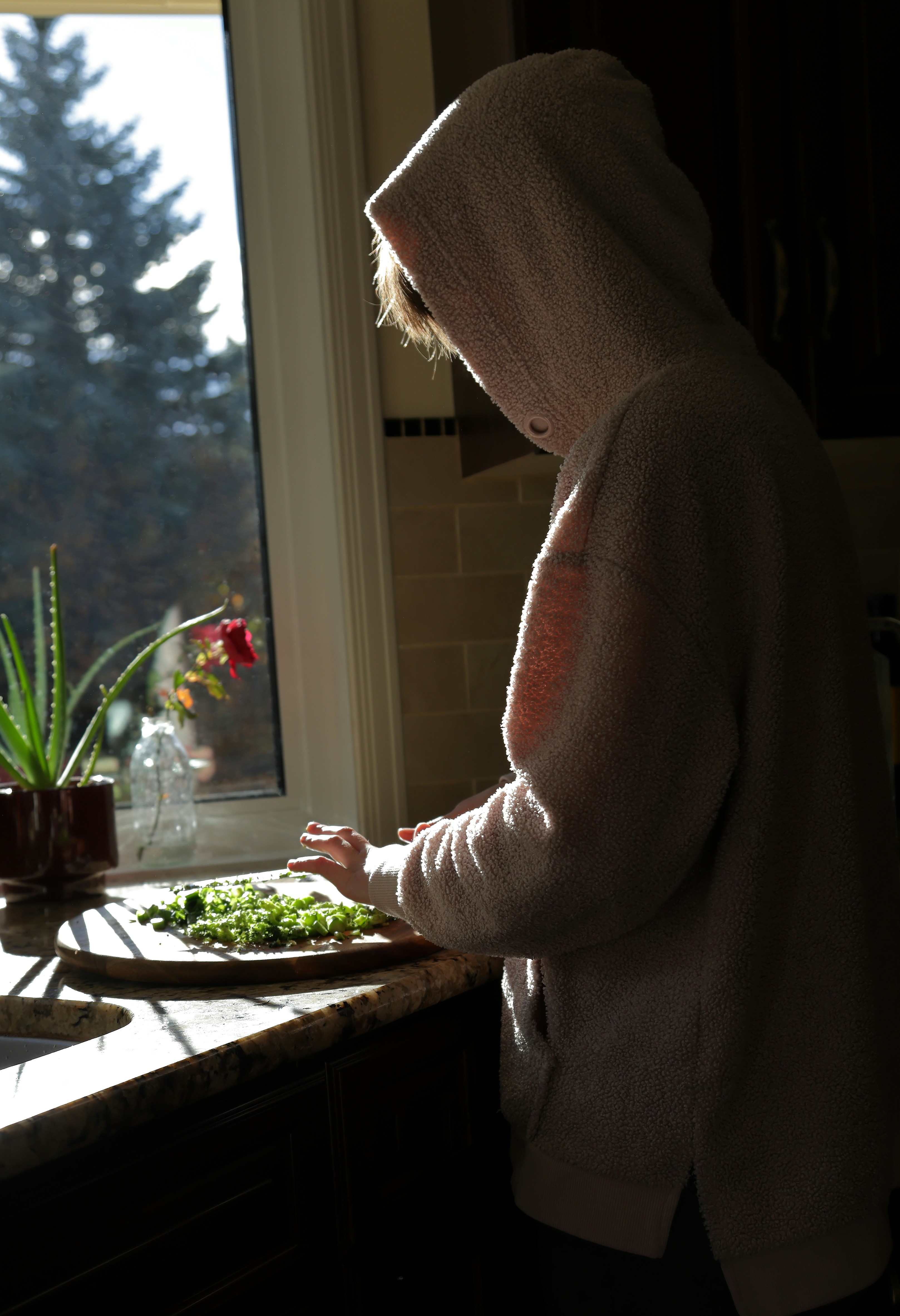A person in a hooded fleece chopping vegetables on a wooden board in a sunlit kitchen