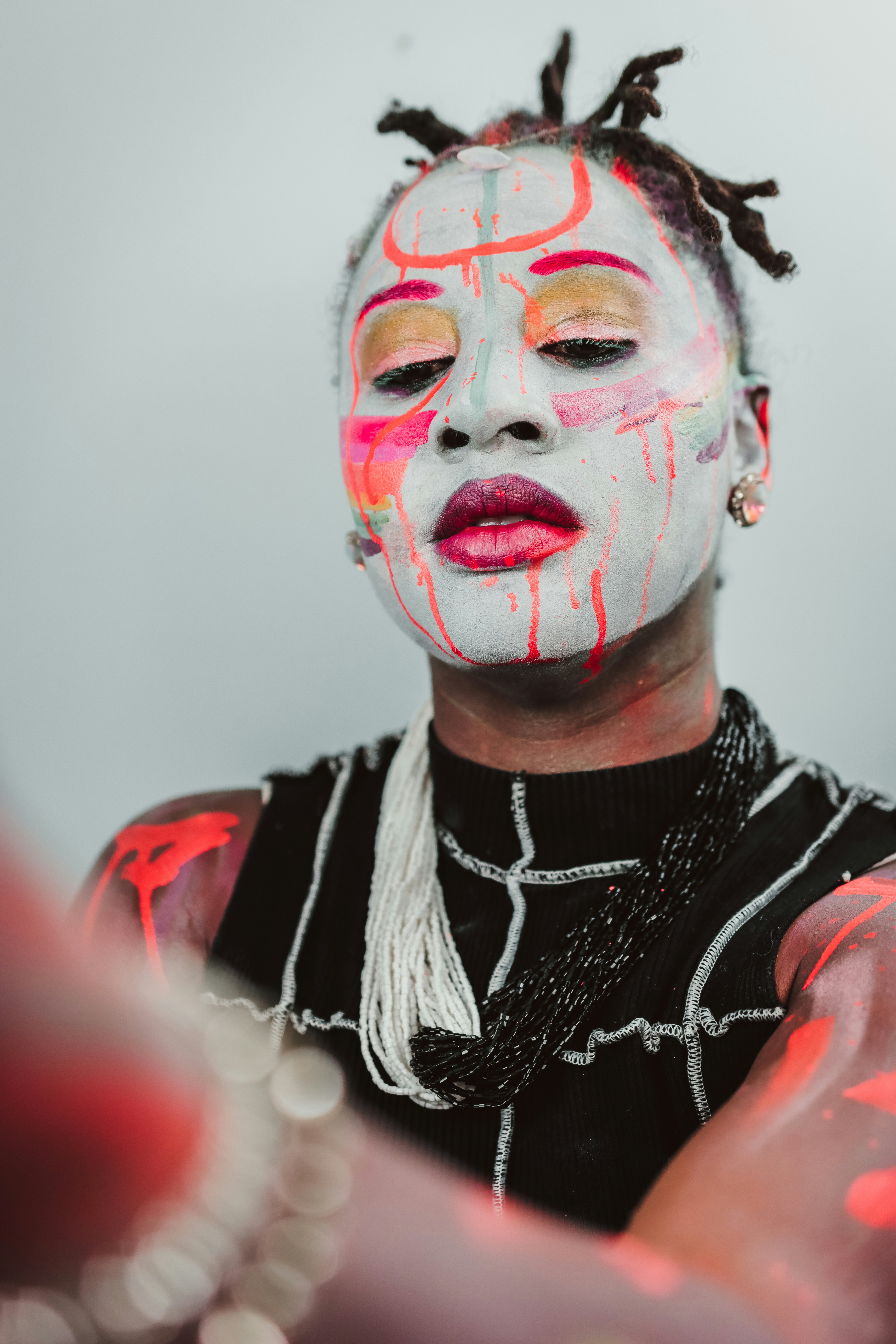 A person with white face paint and neon red markings wearing a black top and beaded necklaces